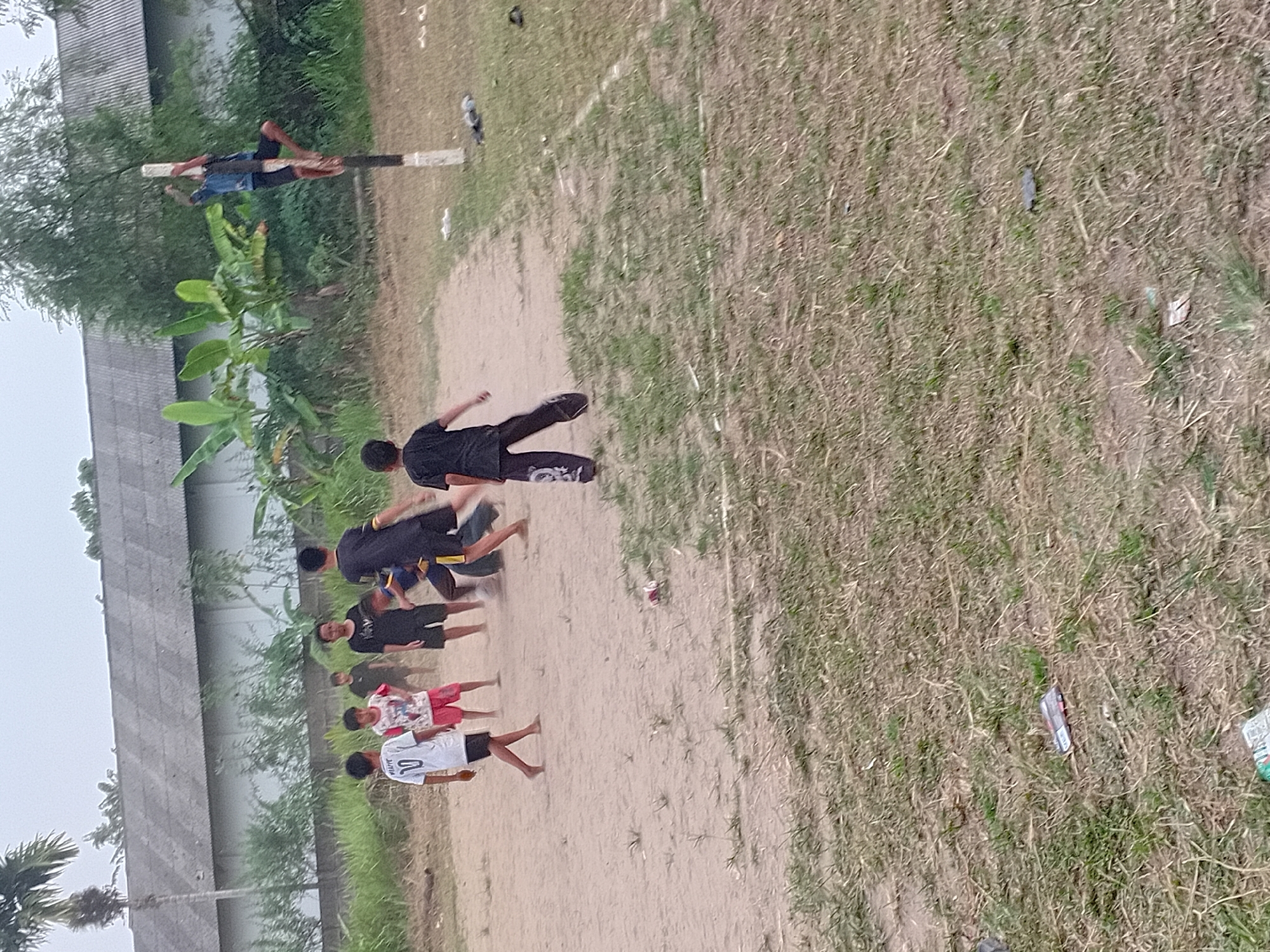A group of young boys are playing soccer on a dusty field in Jambi, Indonesia. One boy is climbing a wooden pole while others are engaged in a soccer game. In the background, there is a building with a gray roof and lush green vegetation. The field is covered in dirt with patches of dry grass. Scattered on the ground are a few discarded items, including a red can and a white plastic bag. The sky is overcast, creating a soft, diffused light.