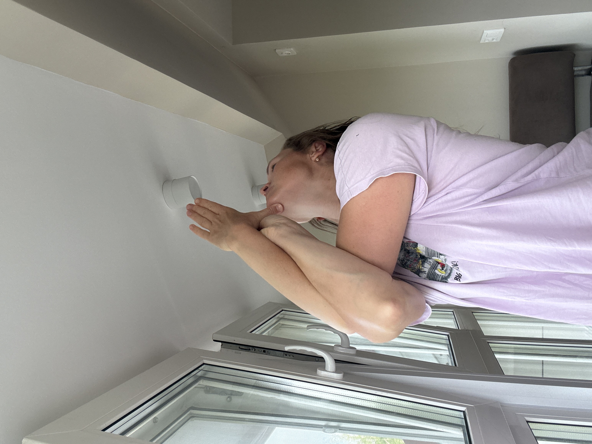 A person is kissing a white cylindrical air vent attached to a white wall. The person is leaning against a partially open white-framed window. The location is likely indoors. The city of Chaikovsky, Russia is not visibly discernible in this image.