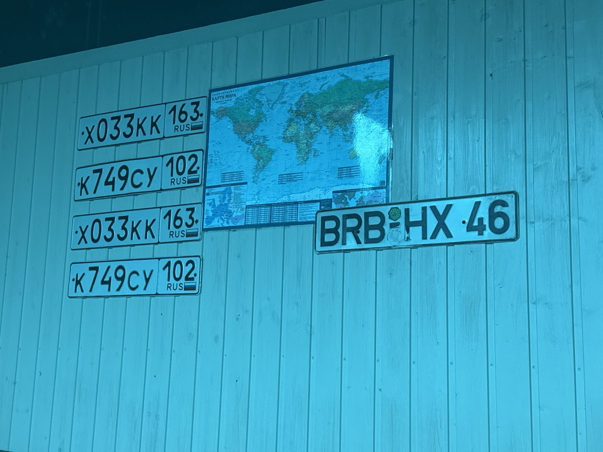 This media file shows a wall covered in wooden planks, painted in a bright blue hue. Affixed to the wall are several objects. Prominently displayed is a large world map, titled "КАРТА МИРА" (World Map) in Russian text. Below the map, and also to its left, are several license plates. Four license plates are stacked vertically on the left side of the image, each appearing to be Russian. Three of these plates display the same characters: "x033KK" followed by a number and "RUS". The fourth plate has different characters: "K749cy" followed by a number and "RUS". To the right of the world map, a single, larger license plate is visible, displaying the characters "BRB HX 46". The lighting suggests an indoor setting, possibly during the day due to the even illumination. There are no people visible in the image, and the overall impression is one of display or collection of these items. The blue tint of the image might be due to the lighting or a post-processing effect.