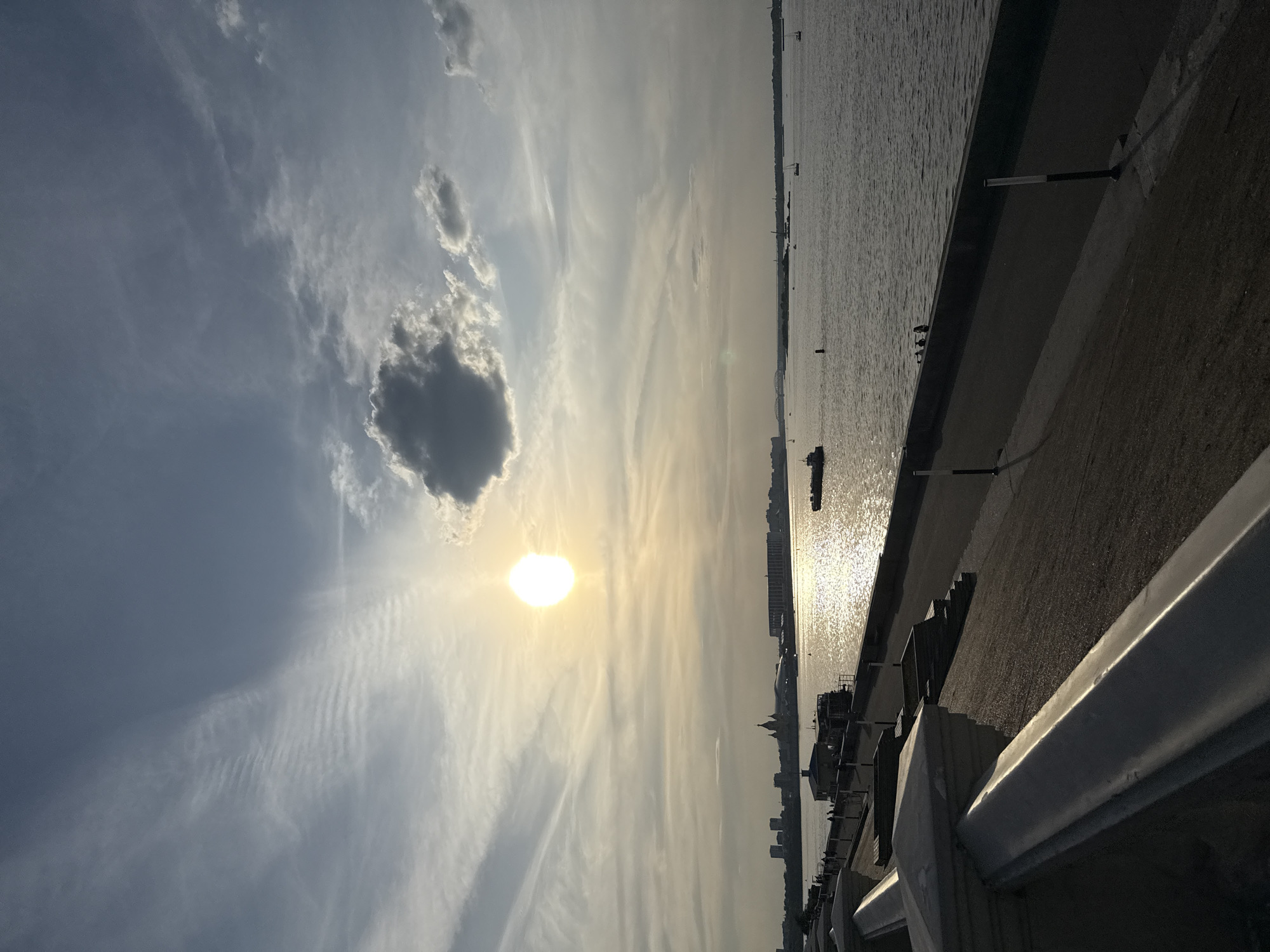 The image captures a serene riverside scene in Nizhny Novgorod, Russia, during what appears to be late afternoon or early evening. The sun, a bright, diffused orb, is low in the sky, casting a warm, golden glow across the water and illuminating the wispy cirrus clouds that streak across the expanse.
The foreground is dominated by a paved embankment, with a white railing running along its edge, suggesting a public promenade or viewing area. Beyond the railing, the wide Volga River stretches out, its surface shimmering with reflected sunlight. A few small boats are visible on the water, hinting at recreational activity or local transport.
In the mid-ground, the distant cityscape of Nizhny Novgorod is silhouetted against the bright sky. Architectural details, including church domes and modern buildings, can be discerned, providing context to the location. The overall atmosphere is peaceful and tranquil, evoked by the soft light and the vastness of the sky and water. The weather appears clear, with the clouds adding texture and visual interest to the sky. There is no visible text in the image.
