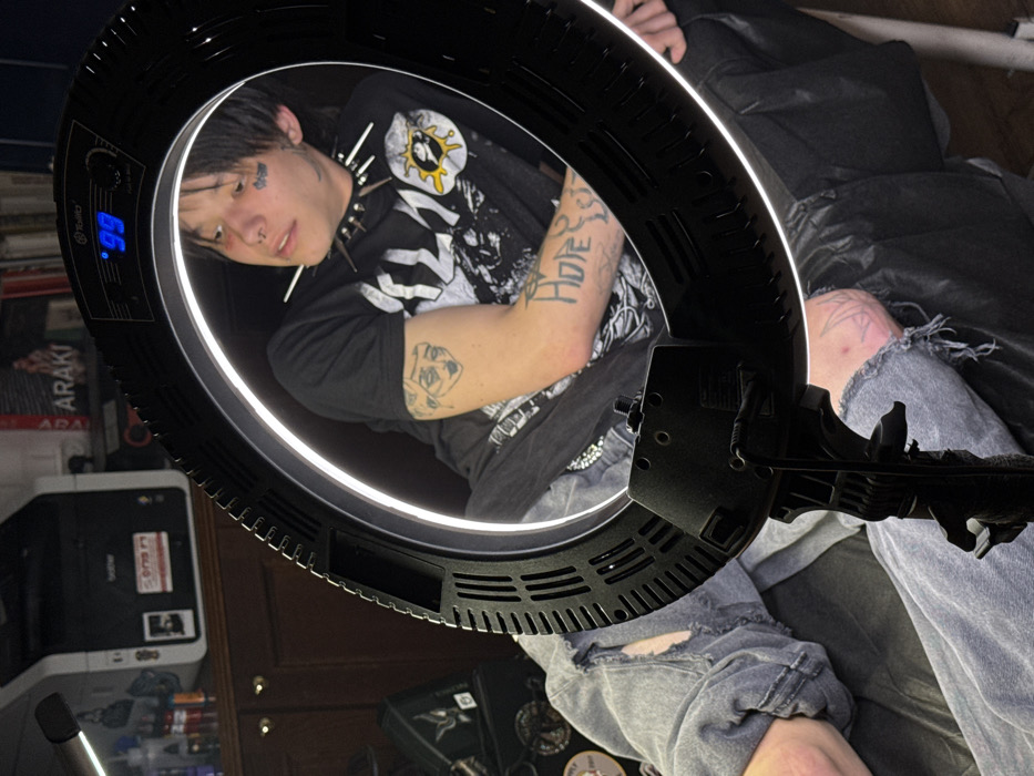 A person with multiple tattoos, including one on their face and arm reading "Hope," is positioned within a black ring light. They wear a t-shirt with a graphic print, a studded collar, and ripped grey jeans. The background includes a stack of books, a Brother printer, and a dark material draped behind them, suggesting a casual indoor setting.