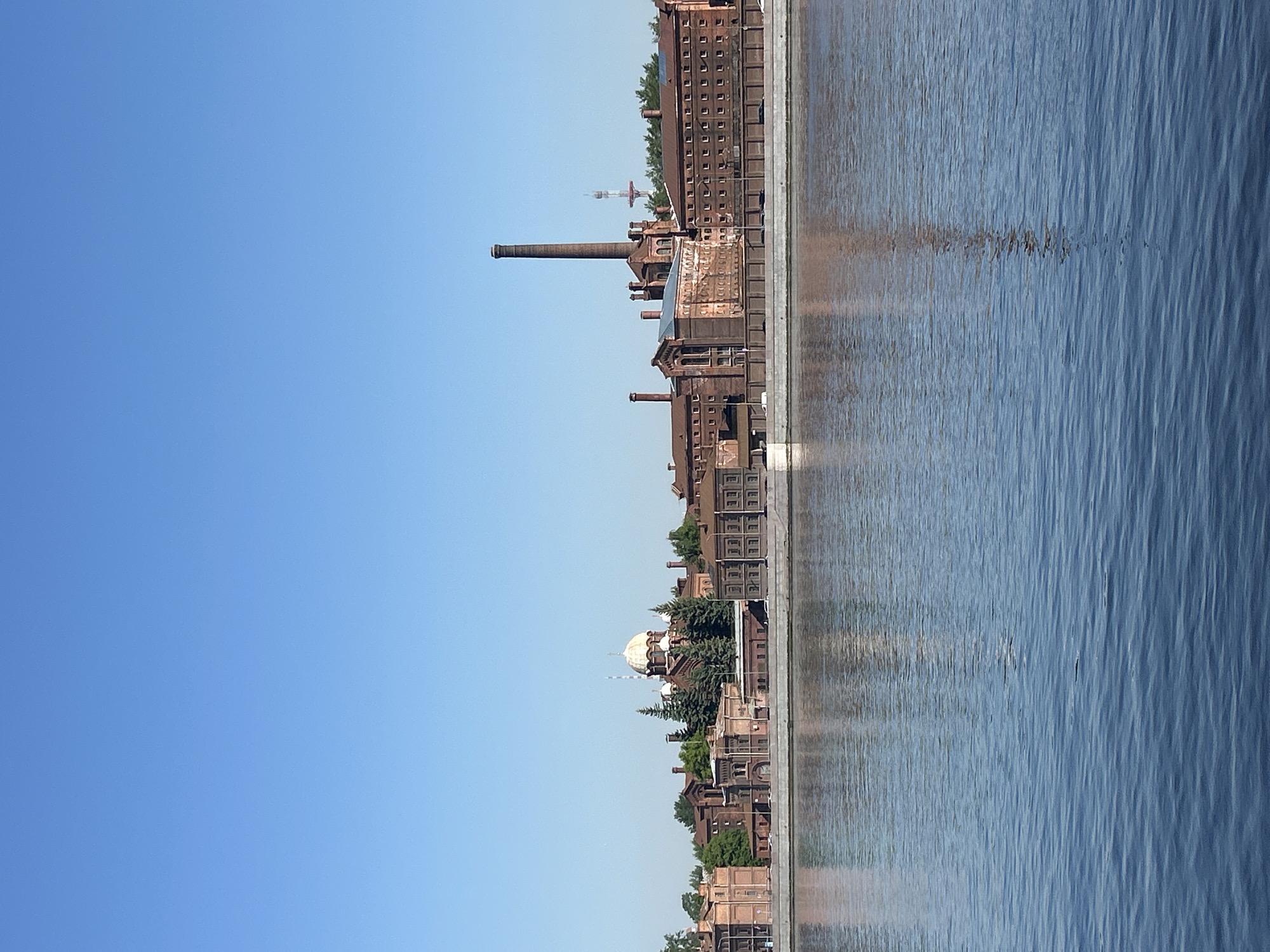 The image captures a wide, panoramic view of a lengthy industrial or historical building complex situated along the bank of a body of water, likely a river or canal, under a bright, clear blue sky.
The upper portion of the frame is dominated by a cloudless blue sky, transitioning subtly from a deeper blue at the top to a lighter shade closer to the horizon.
Along the middle ground, a continuous stretch of reddish-brown brick buildings forms the opposite bank. These structures exhibit architectural characteristics suggesting an older, possibly industrial period, with numerous windows and sturdy construction. Several tall, slender smokestacks or chimneys punctuate the skyline, hinting at the area's past function. Towards the right side of the complex, a distinctive building features a prominent, light-colored dome, partially obscured by green trees. In the far distance, behind the buildings, a tall, thin tower is faintly visible.
The lower portion of the image shows the calm surface of the water, which reflects the clear sky and some of the structures on the bank. The water displays gentle ripples and a deep blue-grey hue.
No people or explicit activity are visible, lending a quiet and static quality to the scene. The bright, even lighting indicates it is daytime, likely a sunny morning or afternoon. There is no visible text in the image. The setting suggests an established urban or semi-urban waterfront area, possibly with a rich industrial history.
