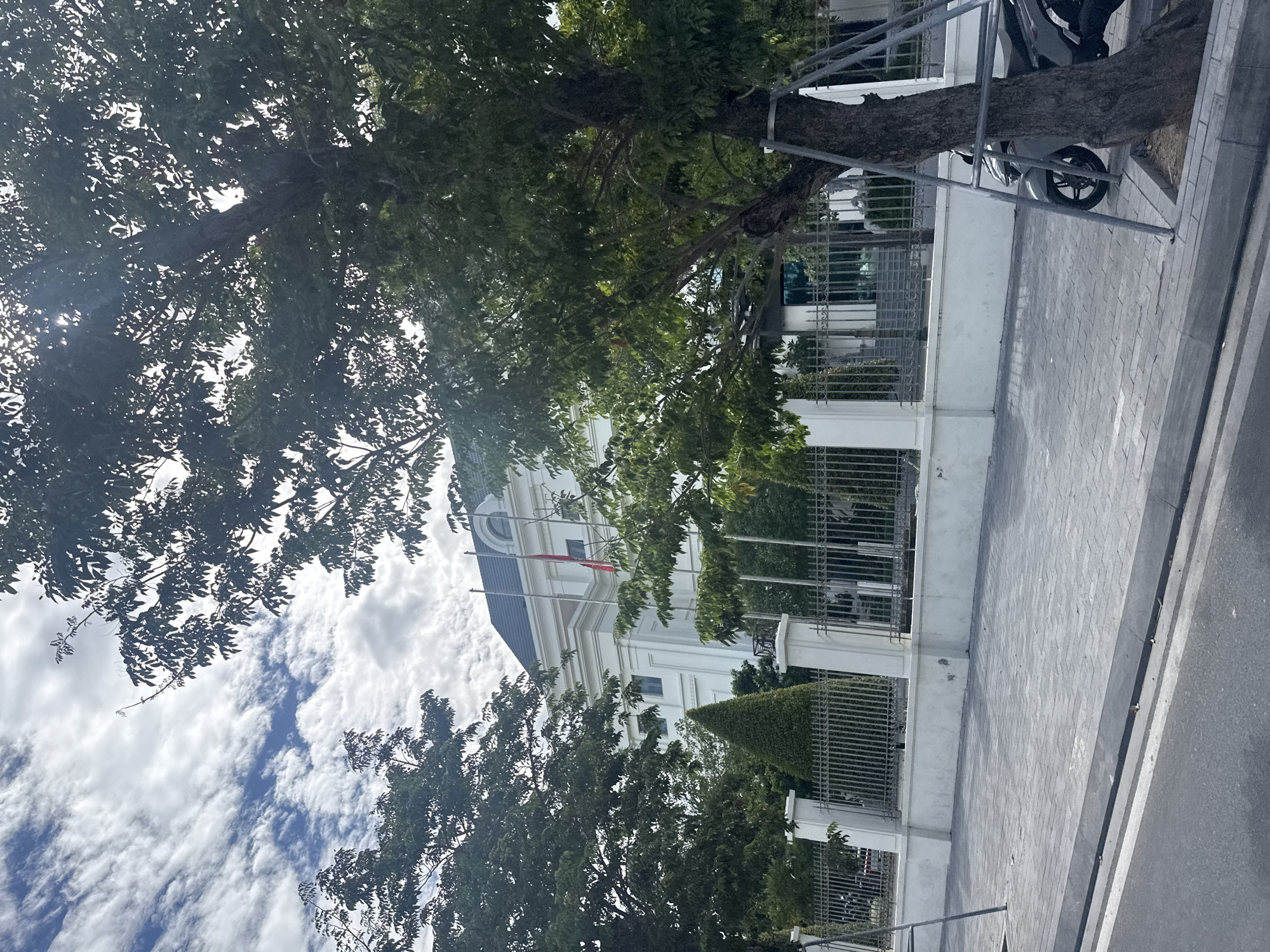 The image captures a street view in Nha Trang, Vietnam, likely on a cloudy day, with a large white building prominently featured behind a decorative fence and lush green trees. The building appears to be modern, with a dark-tiled roof and classical architectural elements, including columns and a rounded window on the upper floor. Two red flags are visible on flagpoles in front of the building.
In the foreground, a paved sidewalk runs parallel to the street. A mature tree with dense foliage stands to the right, its trunk supported by metal bracing. Part of a gray scooter is visible next to the tree, parked on the sidewalk. The street itself is paved, with a curb separating it from the sidewalk. The sky is filled with dramatic, wispy clouds, suggesting dynamic weather conditions. The overall impression is one of a well-maintained urban environment, possibly a government building or an institutional complex, given the flags and formal architecture.
