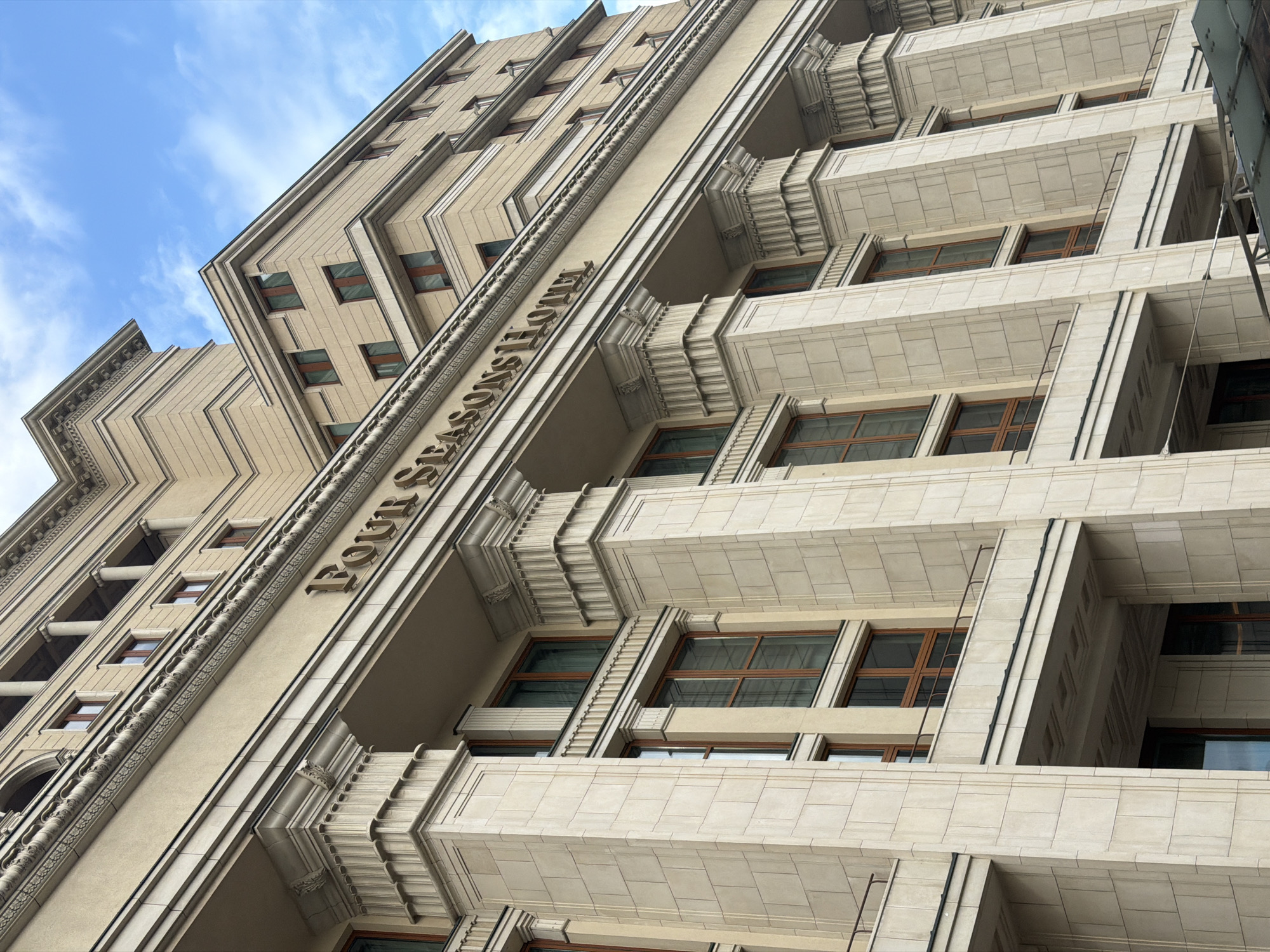 The image shows the Four Seasons Hotel in Moscow, Russia, viewed from a low angle. The hotel's facade is a light beige color with a mix of Art Deco and classical architectural details. The "Four Seasons Hotel" sign is visible on one of the building's sections. The windows are brown and rectangular. The sky is blue with some clouds. A ladder is on one part of the building.