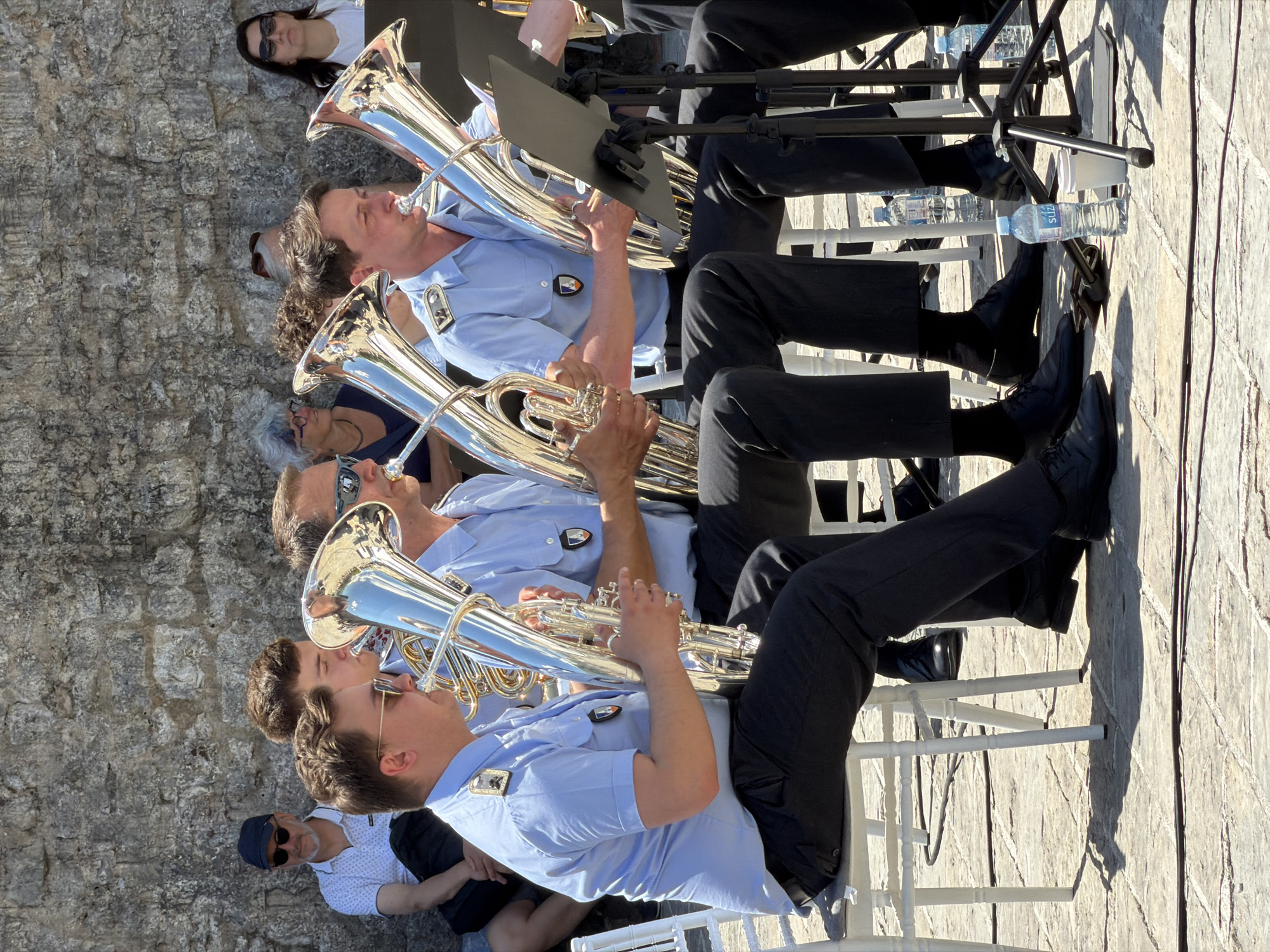 The image depicts a group of musicians seated and playing tubas in Boreti, Montenegro. They are wearing light blue shirts and black pants, possibly uniforms. Each musician is playing a silver tuba. They are seated in a row of white chairs. Behind them is a stone wall. In the foreground are a few seated spectators. There is a water bottle beneath a music stand.