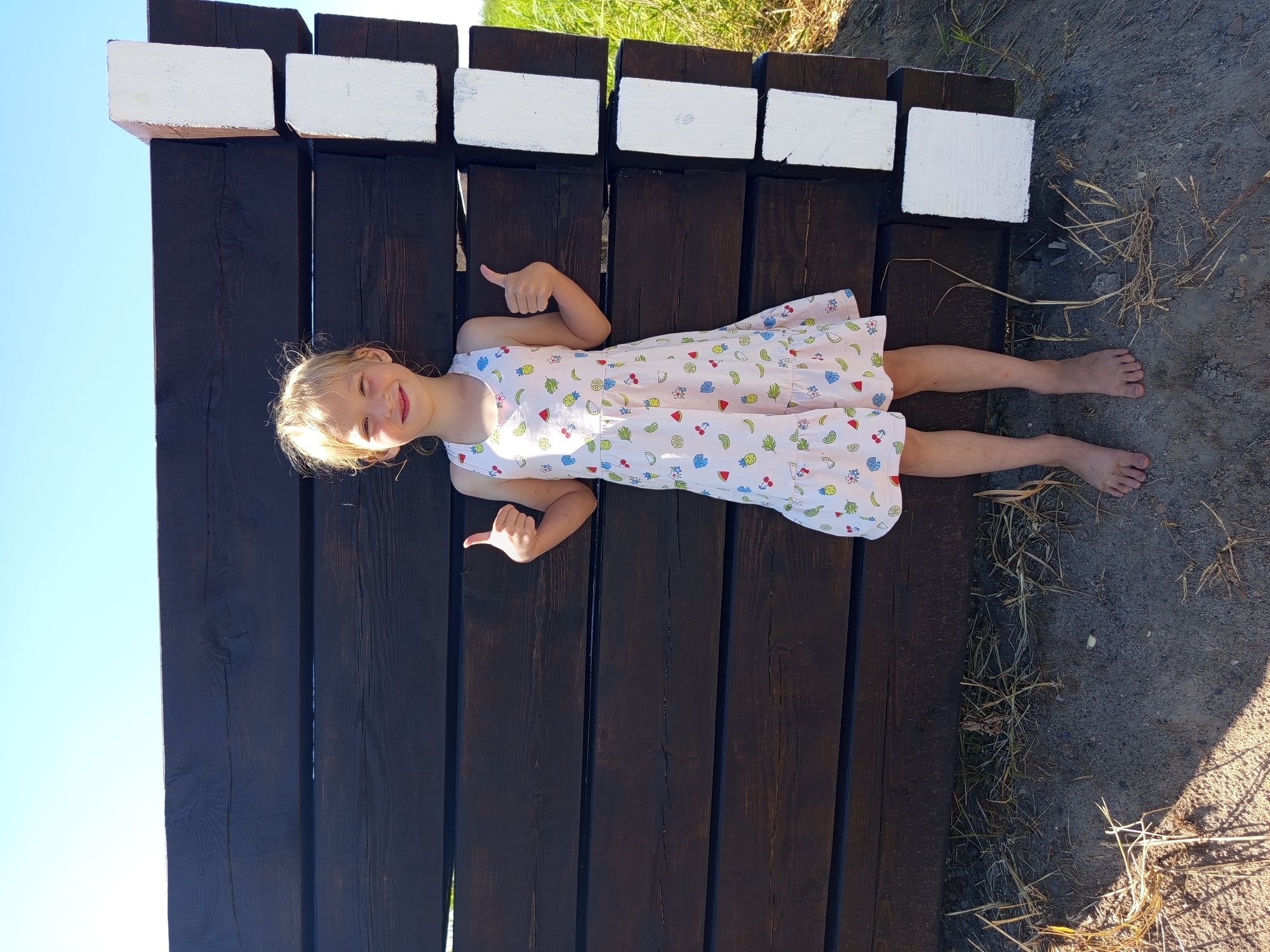 In this media file, a young girl with blonde hair lies on dark wooden planks, her body oriented horizontally in the frame. She is wearing a light-colored dress adorned with a pattern of fruits such as watermelons, pineapples, and bananas. Her facial expression is cheerful, with a wide smile and closed eyes, suggesting happiness or amusement. She is giving a thumbs-up gesture with her left hand, and her right hand is also raised in a similar gesture. Her feet are bare and rest on a patch of dry, greyish soil with sparse vegetation. Above the wooden planks, several white rectangular blocks are visible. The scene appears to be outdoors, with a clear blue sky occupying the upper left portion of the image and the sunlight casting shadows on the ground, indicating it is daytime. The presence of dry grass and the soil suggest a warm, possibly dry, environment. The wooden structure and the open space hint at an outdoor recreational area or a garden setting. The overall mood conveyed is one of joy and relaxation.