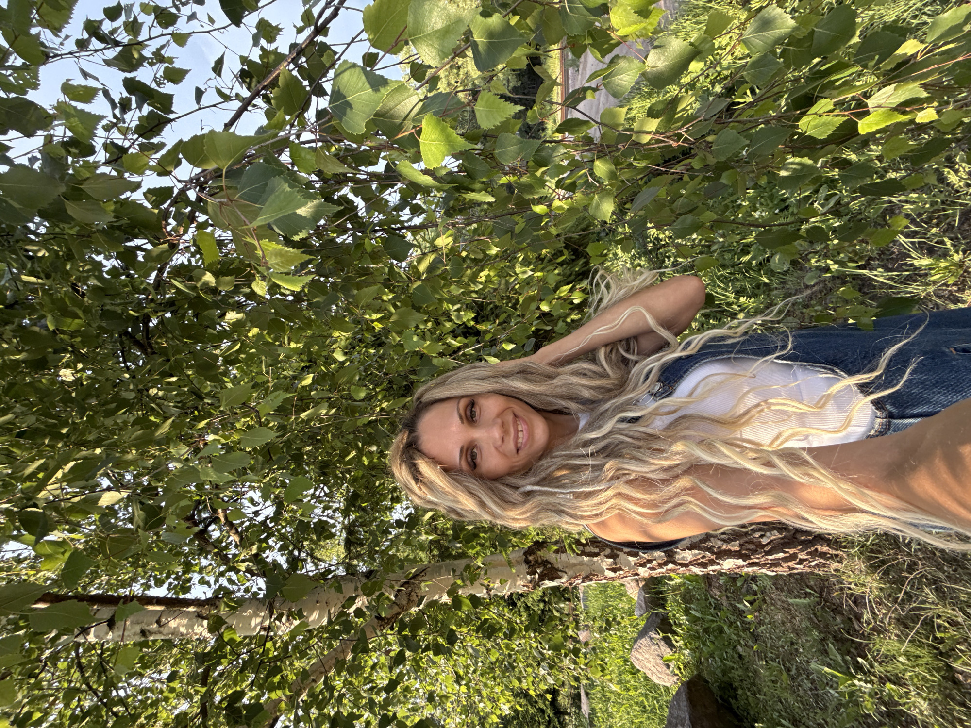 A woman with long blonde hair is standing next to a birch tree in a grassy area. She is wearing a white tank top and denim overalls. The woman is smiling and touching her hair. The background shows more trees and greenery, with a road and some distant trees visible. The lighting suggests it is late afternoon. The location is identified as Rusanovo, Russia.