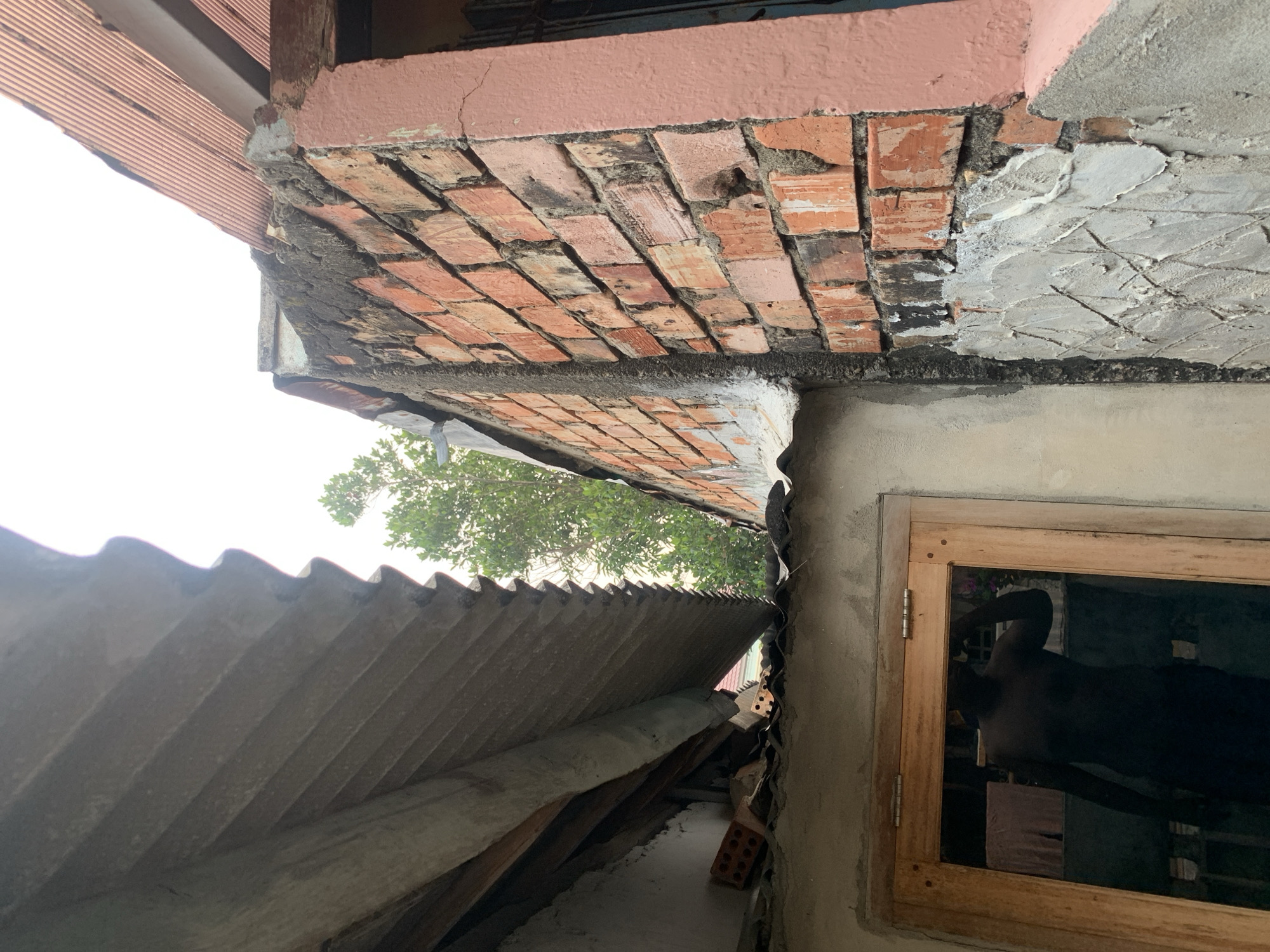 The image depicts an outdoor scene with partially constructed or weathered buildings. The foreground is dominated by a corrugated, light-colored roof section angled downwards. To the right, a brick wall, constructed from reddish-orange bricks with visible mortar, rises vertically. This wall has areas of rough, unfinished grey concrete patching. Beyond the brick wall, a pink-painted structure is partially visible. Further back, lush green foliage from a tree can be seen peeking through the gaps between the structures, suggesting an outdoor, possibly semi-rural or older urban setting. The sky above is overcast and white, indicating a cloudy day. In the lower left portion of the image, a wooden-framed window is visible, and a dark silhouette of a person taking a photo in a mirror is reflected within its glass. The scene conveys a sense of ongoing construction, disrepair, or a simple, practical living environment. This type of construction and the presence of such greenery are commonly found in residential areas within Palembang, Indonesia, particularly in older neighborhoods or areas undergoing gradual development.