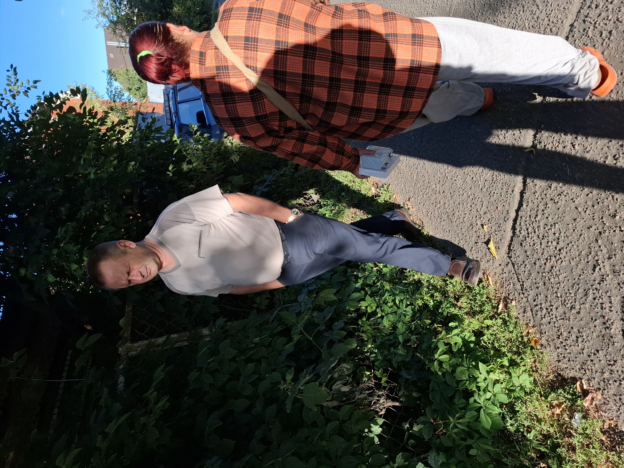 Two individuals walk along a paved path lined with dense green foliage and a chain-link fence on the left. A man in a light-colored short-sleeved shirt and dark pants walks on the left side of the path, his hands in his pockets. A woman wearing an orange and black plaid long-sleeved shirt, light gray pants, and orange slip-on shoes walks on the right side of the path, carrying a white object in her right hand and a bag slung over her left shoulder. A blue vehicle is visible in the background behind the foliage. The scene is illuminated by sunlight, casting distinct shadows on the path.
