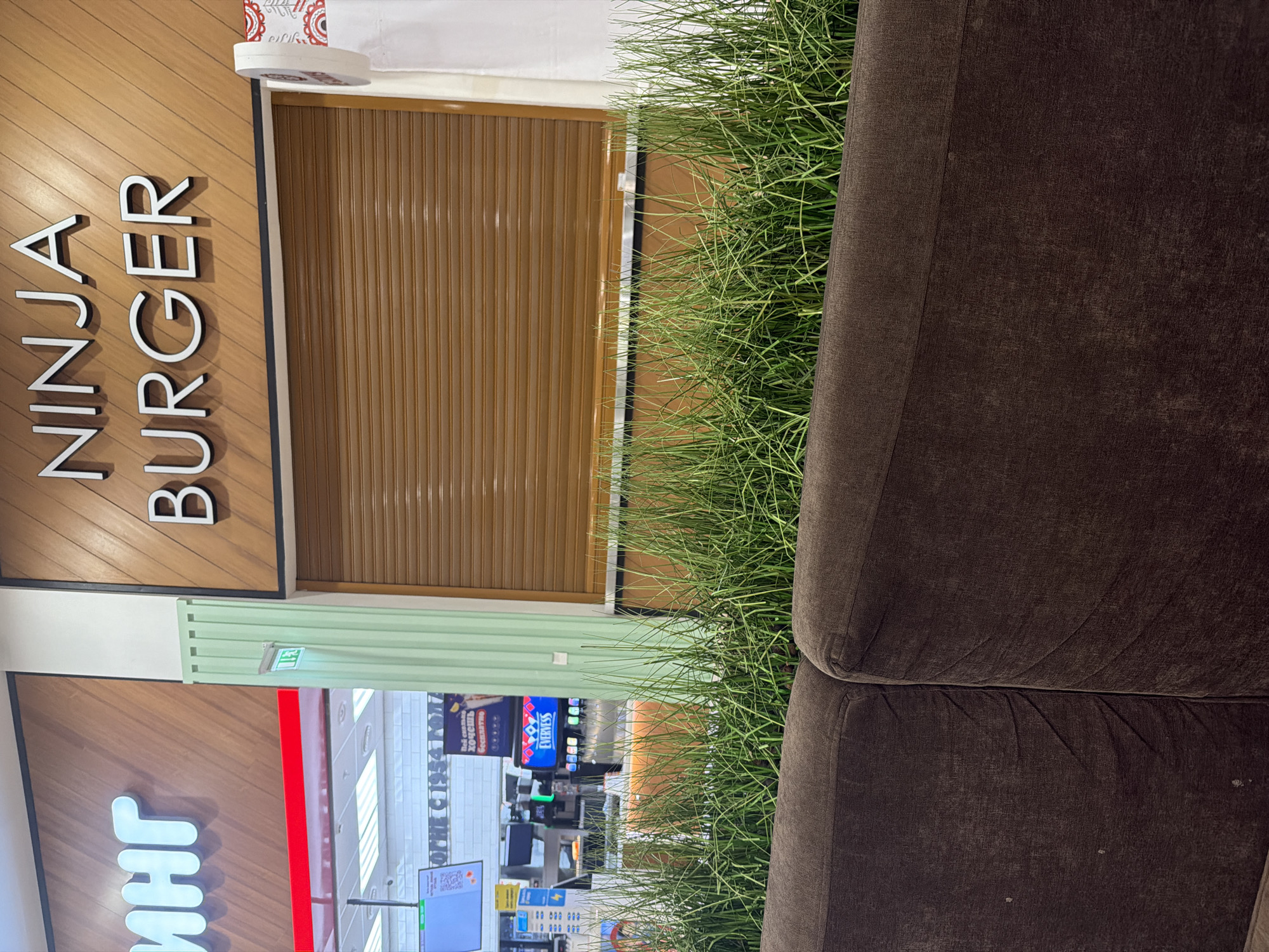 The image shows the exterior of a "NINJA BURGER" restaurant. The sign above the restaurant reads "NINJA BURGER" in large white letters against a wood-paneled background. To the left, a portion of another sign with white letters on a wood-paneled background is visible, with the word "ИНГ" being the most legible. A brown, roller-shutter door is visible in front of the restaurant entrance. In the foreground, a dark brown couch is visible, and behind the couch, a dense arrangement of thin green grass creates a naturalistic barrier. Behind the grass, a slightly blurred view of the restaurant's interior shows a digital menu display with the word "EVERVESS" prominently displayed, along with what appear to be drink dispensers. A green, fluted column is present to the left of the shuttered entrance.
