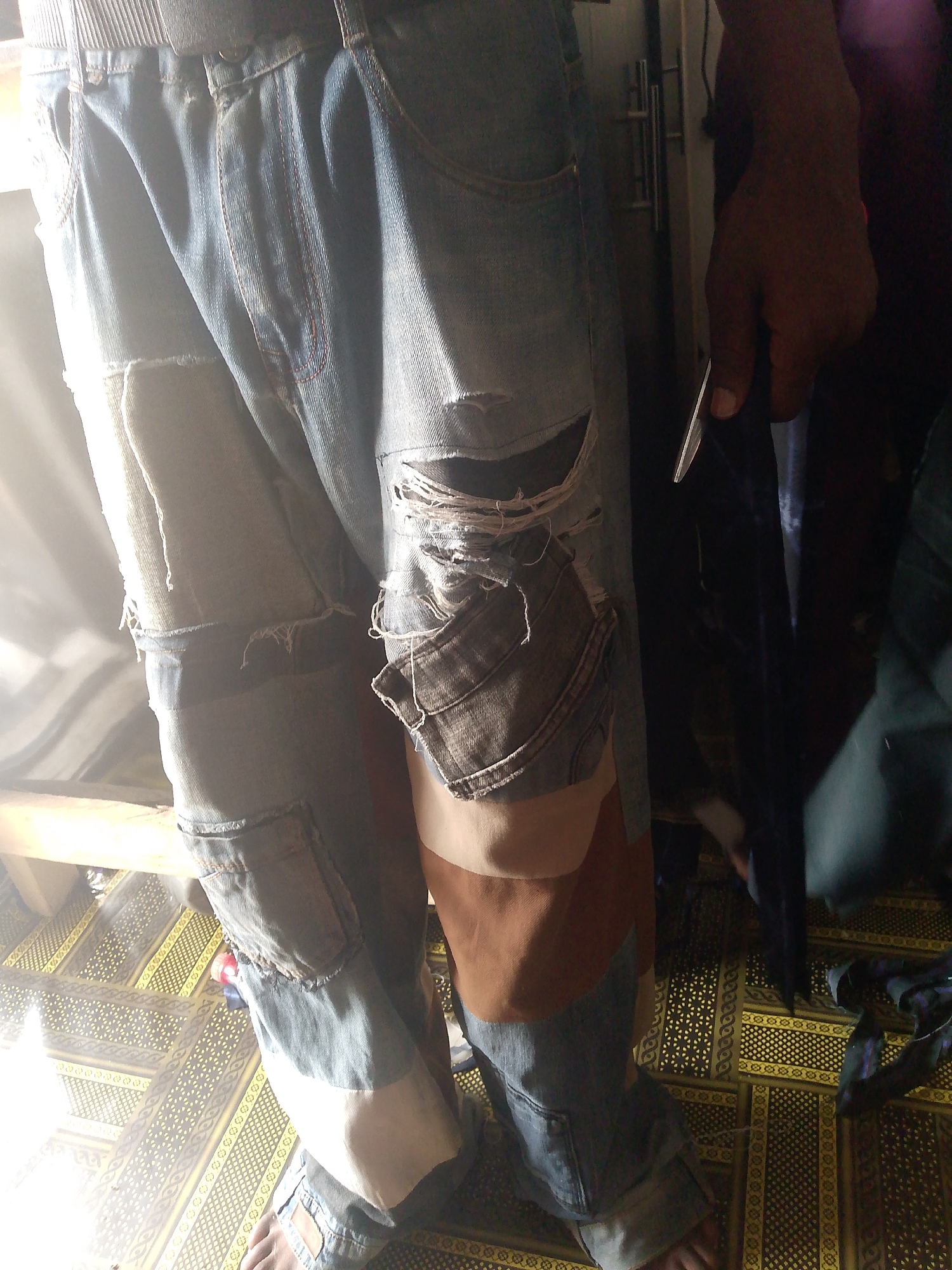 The image shows a person wearing distressed denim jeans with patchwork details, standing indoors. The jeans are blue with lighter blue and brown patches stitched onto them, and feature rips and frayed edges. The person's hand, visible on the right side of the image, is dark-skinned and appears to be holding a dark object. The background is out of focus but suggests an indoor setting with a patterned floor. The scene does not provide any specific visual cues to determine its location in Jalingo, Nigeria.