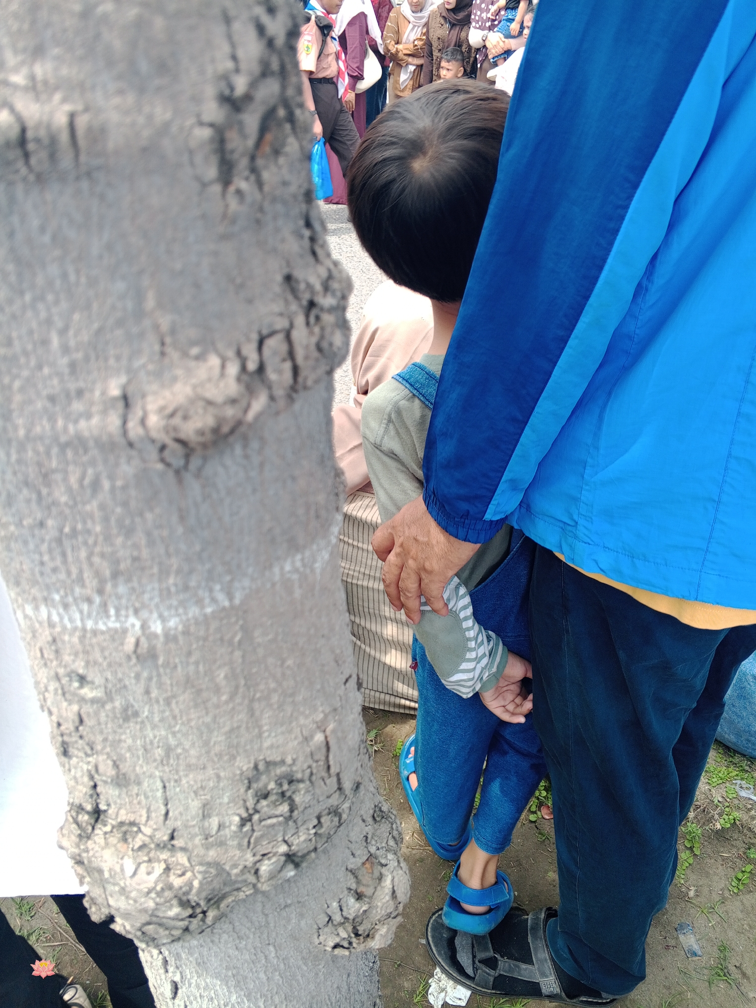 The image captures a close-up view of a young child, with dark hair, wearing denim overalls and a striped long-sleeved shirt, standing next to a tree trunk. The child's left hand is resting on the hip of an adult, whose legs are visible wearing dark pants and a blue jacket. The adult's hand is also resting on the child's arm. In the background, partially obscured by the adult, other individuals are seen, some wearing traditional clothing. The ground appears to be dirt with scattered green vegetation. The context suggests a public gathering, possibly an outdoor event or market, in Lhokseumawe, Indonesia.