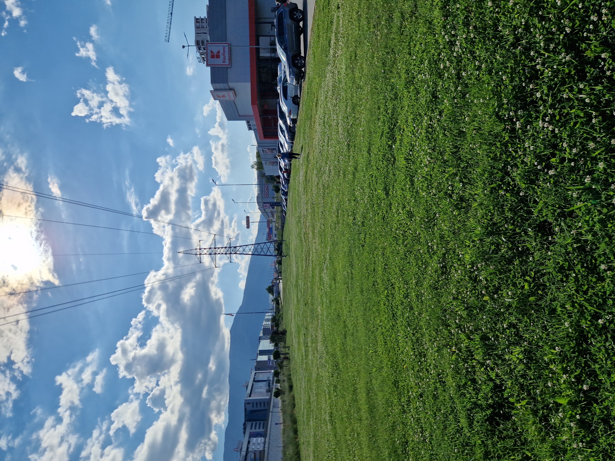 In Sofia, Bulgaria, a wide expanse of lush green grass, speckled with white clover, dominates the foreground of the image. In the middle ground, a row of parked cars lines the edge of a paved area. Beyond the cars, a modern commercial building, identified by a "Kaufland" sign, stands prominently. Further in the background, several other buildings and a construction crane are visible, with mountains forming the backdrop under a bright blue sky filled with scattered white clouds. Overhead, high-voltage power lines stretch across the frame, supported by a tall metal tower. A lone person is walking on the paved area near the parked cars.