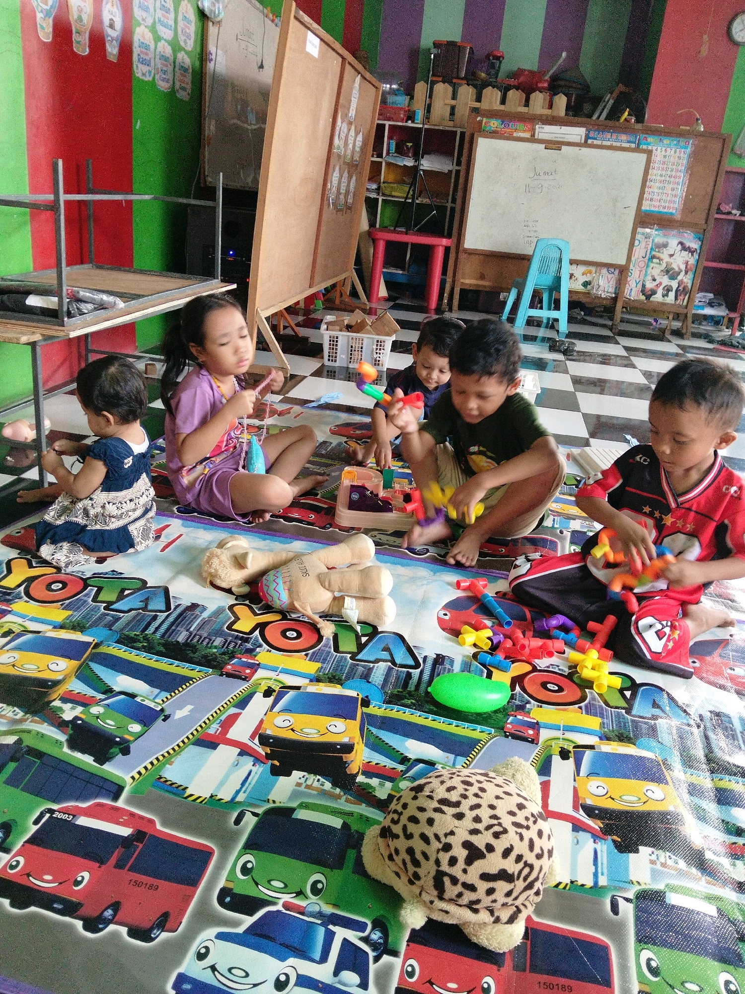In an indoor setting with a black and white checkered floor, several young children are playing on a colorful mat depicting cartoon buses and city scenes. In the foreground, a girl in a purple shirt and shorts sits cross-legged, holding a pink string. To her left, a baby in a blue and white dress sits facing away from the camera. In the center of the image, three boys are playing with colorful building blocks. One boy, wearing a dark green shirt and tan shorts, is focused on the blocks. Another boy, in a red and black jersey, is also engaged with the blocks. A third boy, partially visible behind them, wears a dark blue shirt. Several plush toys, including a tan teddy bear and a spotted turtle, are scattered on the mat. In the background, a whiteboard on an easel is visible, along with shelves containing books and toys. The room appears to be a classroom or play area, with brightly colored walls and educational materials.