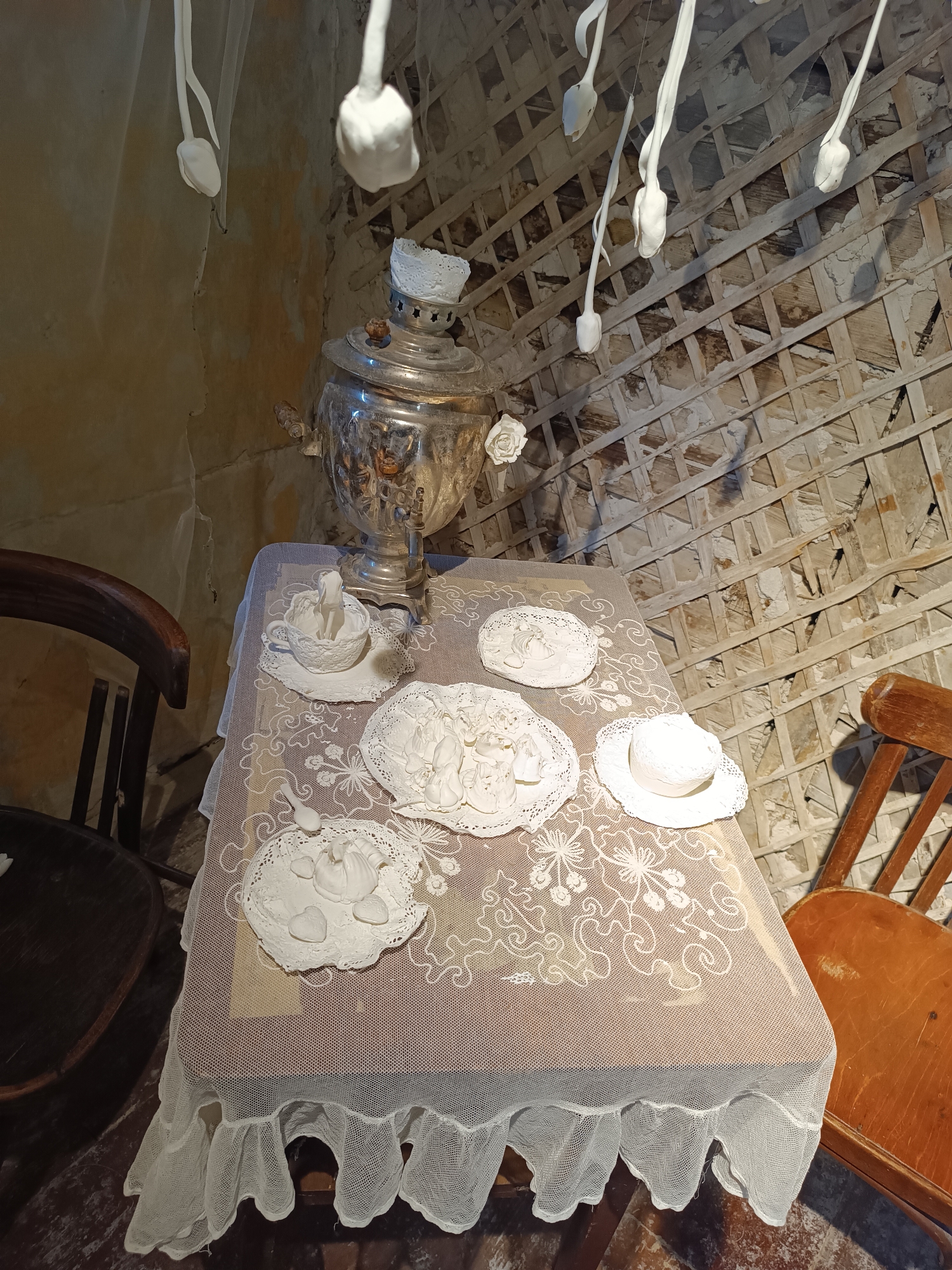 The image depicts a still life arrangement in a room with rough, textured walls and a lattice-like wooden structure on the ceiling. In the center, a table is covered with a light-colored, lacy tablecloth. On the table, there is a silver samovar, a teacup with a white lace doily, and several white sculptural elements that resemble small hats and intricately detailed figures. There are also white decorative roses visible. Two wooden chairs are positioned on either side of the table. Hanging from the ceiling are several white, abstract forms that appear to be made of a soft material, possibly fabric or paper. The overall impression is one of a whimsical or artistic display, possibly set up for a special occasion or exhibition. This scene is located in the "Teehaus" (Tea House) exhibit within the Yelets Museum of Local Lore in Yelets, Russia.