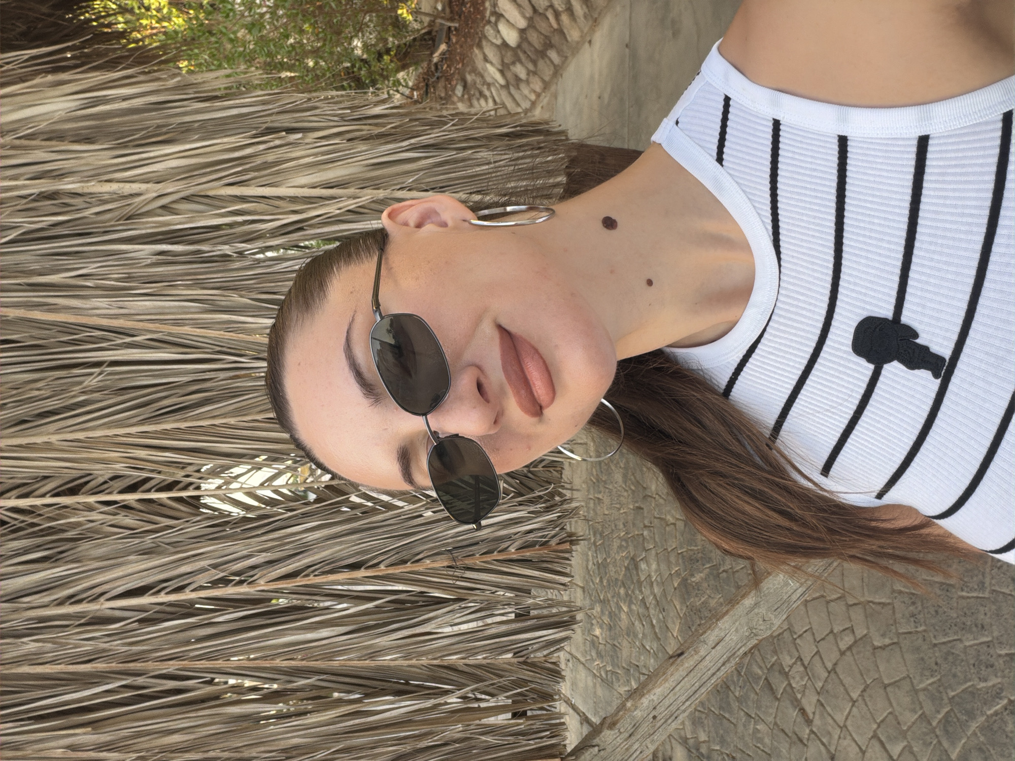 A woman is taking a selfie outdoors. She is wearing sunglasses, hoop earrings, and a white tank top with black stripes. She has long, brown hair and a mole on her neck. Behind her is a wall made of dried palm fronds and a paved walkway. The scene appears to be in a sunny, outdoor location. The user is referencing Sharm El Sheikh, Egypt.