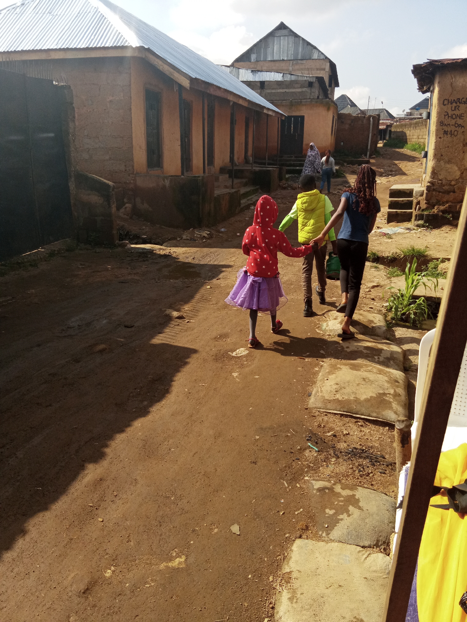 The image depicts a dusty, narrow street in the city of Jos, Nigeria. On the left, a row of buildings constructed with cement blocks and topped with corrugated iron roofs line the street. A large black metal gate is visible in the foreground on the left. Further down the street, on the right, another building made of stone and mud has graffiti on its wall that reads "CHARGE UR PHONE 8AM - 6PM N40". The ground is uneven and appears to be dirt and sand, with some patches of wetness and scattered debris.
In the center of the image, three individuals are walking away from the camera. The closest is a young girl wearing a red hooded jacket with white stars and a purple skirt. She is holding hands with a child in a lime green jacket. Behind them, a young woman with dark braided hair, wearing a dark blue top and black pants, is walking. The children appear to be engaged in conversation or play, their body language suggesting a casual interaction. The overall scene portrays a typical street in a developing urban area, with a mix of residential and commercial structures and evidence of everyday life.