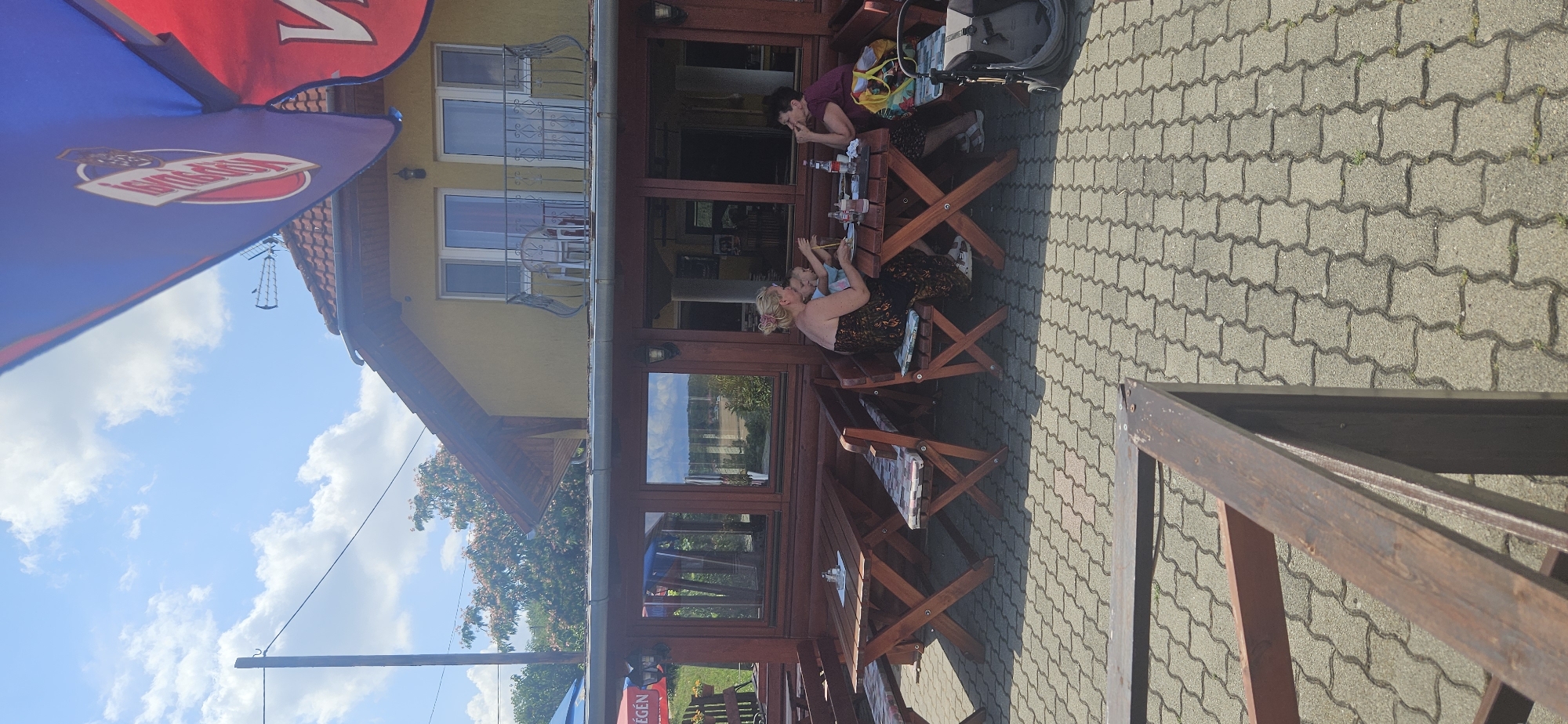 The image depicts an outdoor seating area of a restaurant, likely in Hévíz, Hungary, given the style of the building and the presence of umbrellas with what appear to be Hungarian text. The scene is set on a sunny day with a blue sky and scattered clouds.
In the foreground, a paved area with interlocking grey stones is visible, with a wooden railing in the lower left corner. Further back, several wooden picnic-style tables and benches are arranged. Some of the benches have patterned cushions.
At one of the tables, three individuals are seated. On the left, a woman with blonde hair tied up in a bun is wearing a strapless, patterned dress. She is interacting with a young child, who is wearing a blue top and appears to be eating or playing with food. To the right of them sits another woman with dark hair, wearing a purple top. She has her hand near her face and appears to be looking away from the table.
On the table, there are bottles of drinks, glasses, and possibly some food items. A bag is placed on the bench next to the woman in the purple top, and a stroller or backpack is visible to the far right.
Behind the seating area is a wooden-framed structure with large windows, reflecting the sky and trees. Above this structure is a yellow building with a tiled roof. The building has windows and a balcony with a decorative railing on the upper floor. Two large umbrellas, one predominantly blue and red, are positioned above the outdoor seating area, providing shade. One umbrella features a logo with text that suggests a beverage brand.
The overall impression is of a casual outdoor dining or relaxation spot.