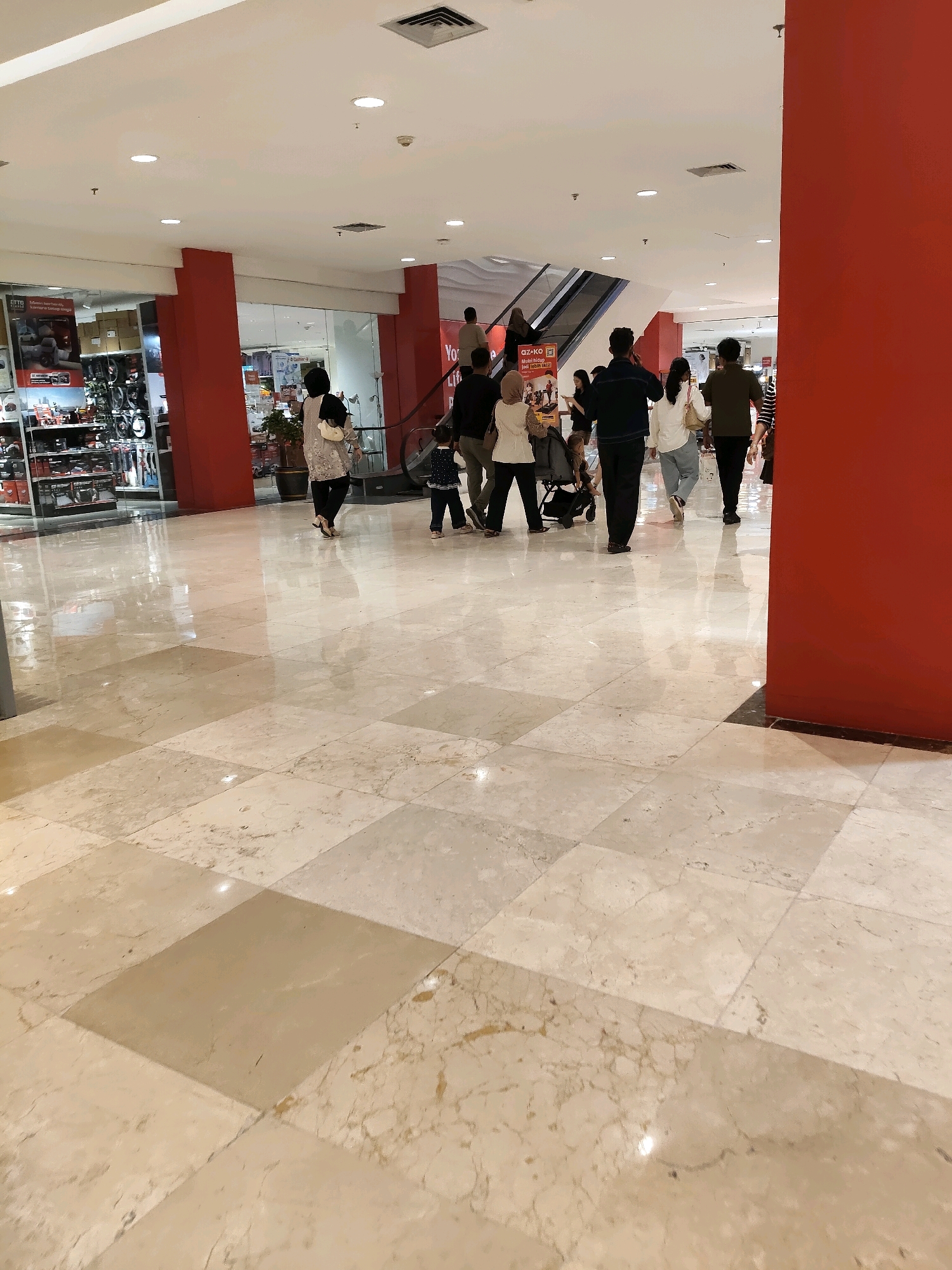 The image depicts the interior of a brightly lit shopping mall in Medan, Indonesia. The floor is paved with large, polished marble tiles in beige and cream tones, reflecting the overhead lights. People are walking through the mall, some in groups, others individually. In the background, there are escalators and shop fronts, suggesting a commercial setting. One shop display on the left shows what appears to be automotive parts or accessories. A prominent red pillar divides the space. The ceiling features recessed lighting and air vents. The scene captures a typical day in a public commercial space.