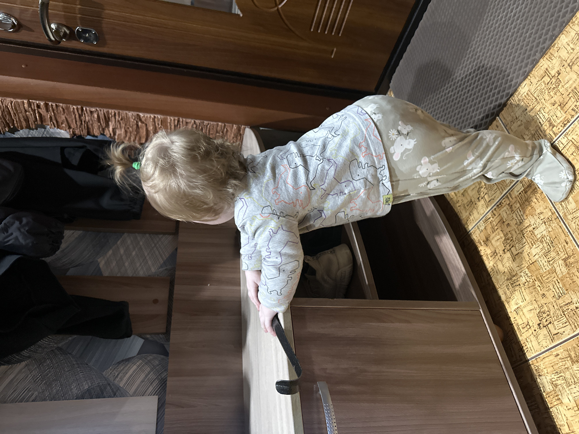 A blond-haired toddler, wearing a light grey long-sleeved shirt with a dinosaur pattern and light green pajamas adorned with mice and flowers, stands in a hallway. The child is facing away from the camera, looking towards a wooden cabinet and a brown door. The toddler's left hand rests on the edge of the cabinet, which has a wooden front and a metal handle. A black metal object, possibly a shoehorn, is hooked onto the cabinet's edge. Inside a shelf of the cabinet, a pair of white sneakers is visible. The floor is covered in light brown square tiles with a wood-like pattern. To the right of the cabinet, a brown wooden door with a silver handle and lock is present. The overall environment appears to be a home interior, likely in an apartment or house. The image does not contain specific geographical markers to pinpoint a location within Sayanogorsk, Russia, beyond it being an indoor residential setting.