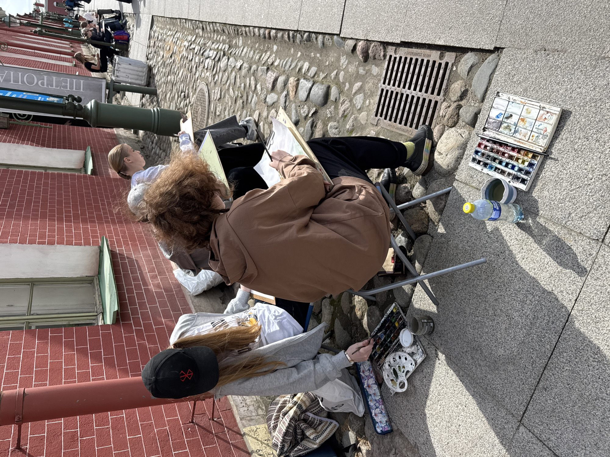 Several individuals are engaged in plein air painting or sketching on a sunny day in what appears to be Saint Petersburg, Russia. The scene is captured from an elevated perspective, looking down and slightly across.
In the foreground and mid-ground, three young people are seated on portable chairs, focused on their artwork. One person, with curly brown hair and wearing a brown hoodie, is actively painting on a canvas or sketchbook held on a clipboard. To their left, another individual with blonde hair, wearing a light-colored top, is also drawing or painting. A third person, with long blonde hair and wearing a grey hoodie and a black baseball cap with a red symbol (possibly a cross or similar design), is painting with watercolors, a palette and water cup visible beside them on the grey pavement.
Art supplies are scattered on the ground near the artists: a large watercolor palette with multiple pans of paint, another smaller palette, water bottles (one clearly branded "BON AQUA"), and plastic cups for water. The ground is a mix of light grey granite pavement and cobblestones, with a metal drain cover visible. Strong shadows cast by the artists and nearby objects indicate bright sunlight, suggesting midday or early afternoon.
To the left, a tall, dark green pole supports a sign with white text, partially visible, reading "ПЕТРОПА" (likely part of "Петропавловская крепость" - Peter and Paul Fortress, given the context). Behind this, a red brick building with white window frames and green trim forms the backdrop. Other people can be seen further down the street, some seated, others walking, suggesting a public area. The overall atmosphere is calm and focused, with the artists absorbed in their creative activity outdoors.