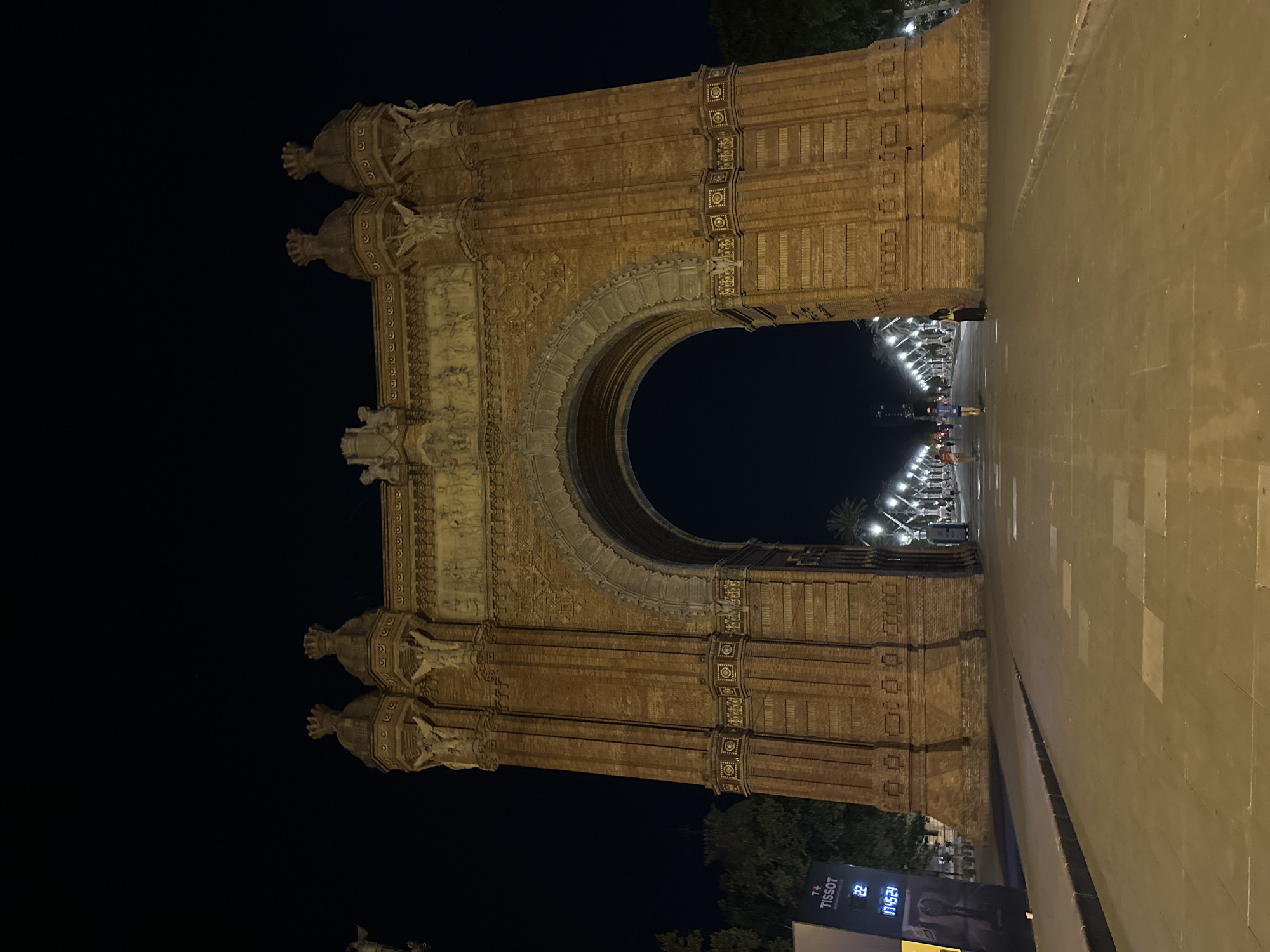 The image depicts the Arc de Triomf in Barcelona, Spain, at night. The monument, constructed from red brick, is illuminated and stands prominently against a dark sky. The archway is wide and tall, with detailed carvings and sculptures adorning its facade. Beyond the arch, a tree-lined avenue extends into the distance, with streetlights casting a warm glow. Several individuals are visible walking along the avenue, some appearing to be jogging. To the left of the arch, a large advertisement display features the Tissot logo and digital readouts, including a time display of "04:524." The ground in front of the arch is paved with large stone slabs.