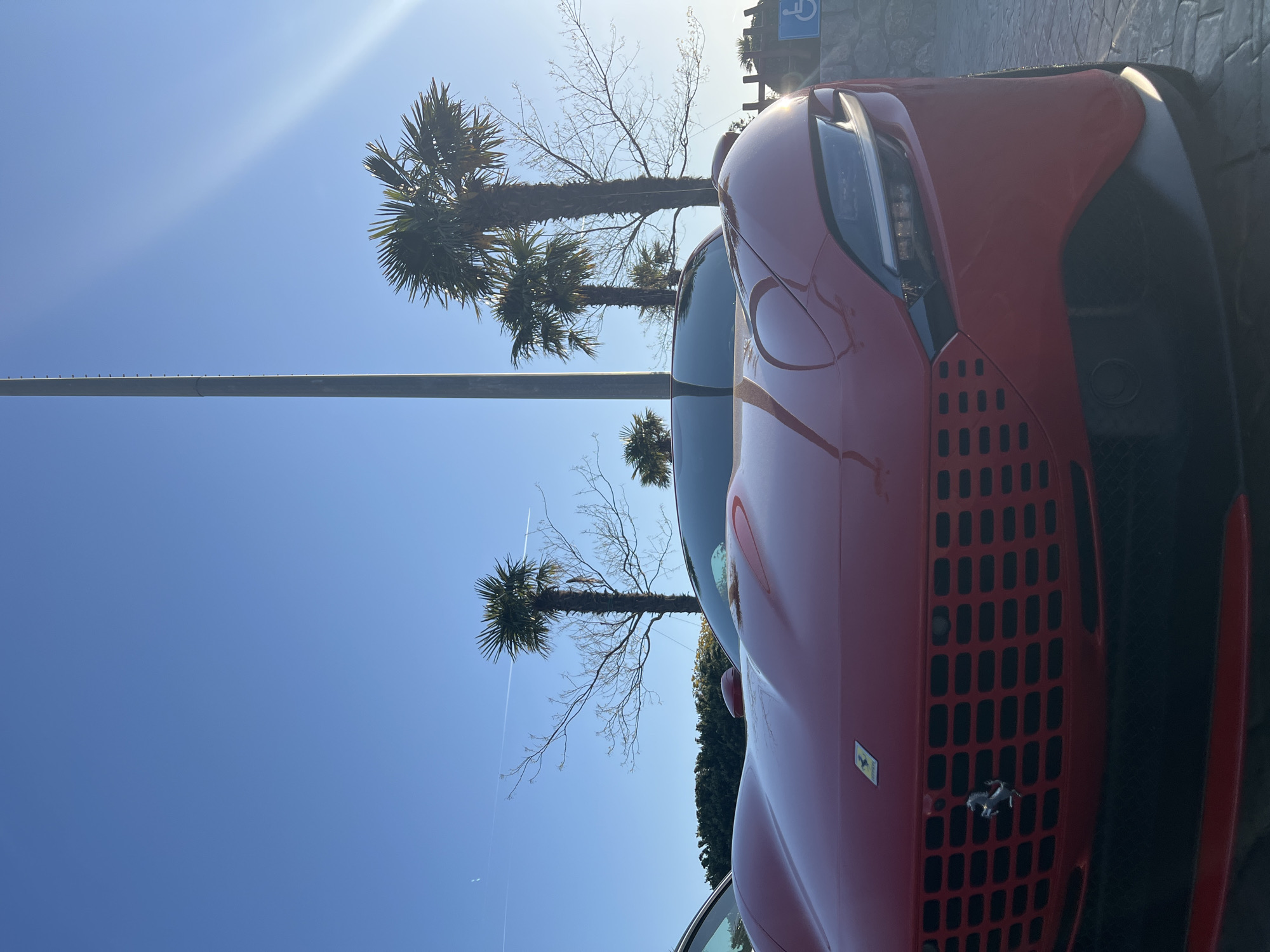 A red Ferrari is parked adjacent to a palm tree-lined street in Kovačko Polje, Montenegro. The scene is bright with a clear blue sky. A long, horizontal support structure runs parallel to the street. The front of the car is prominently displayed, including the grill and the Ferrari emblem. The photo is taken from a low angle, emphasizing the car and the sky.