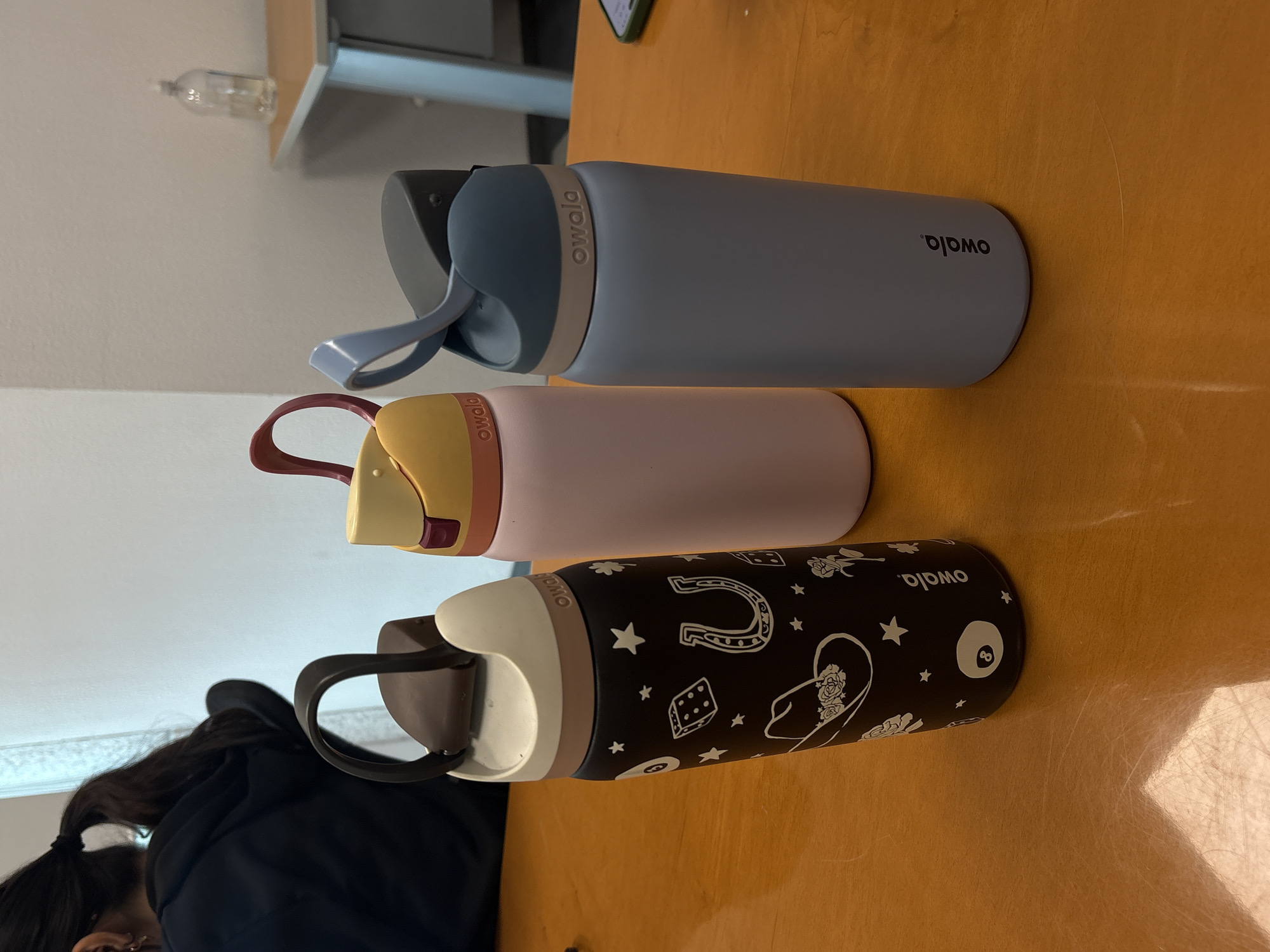 The image shows three Owala water bottles laying horizontally on a yellow table. From top to bottom, the bottles are light blue, pink, and black with western-themed designs. The background shows a blurry view of a shelf and a person. The location, Puebla, Mexico is not discernible.