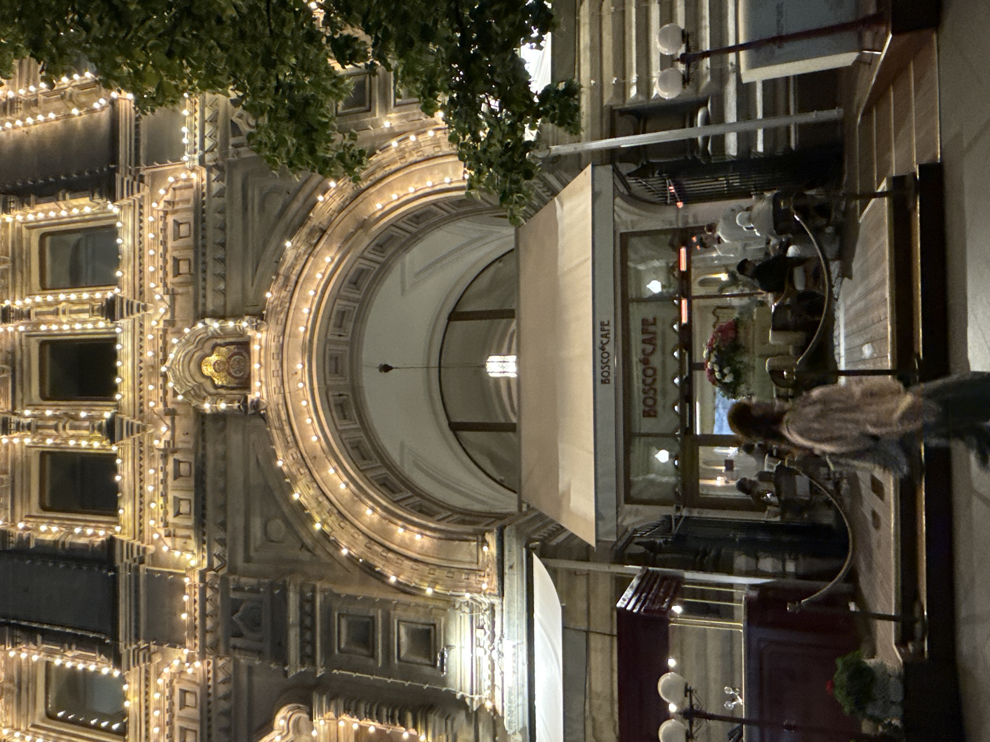 This outdoor shot at night features a building facade with a large arched entrance, decorated with strings of lights. To the right, there's a "Bosco Cafe" with tables and chairs set up outside, some of which are occupied by people. A person is standing in the foreground, near the outdoor seating. The scene appears to be located in Moscow, Russia, given the cafe's presence at a well-known location.