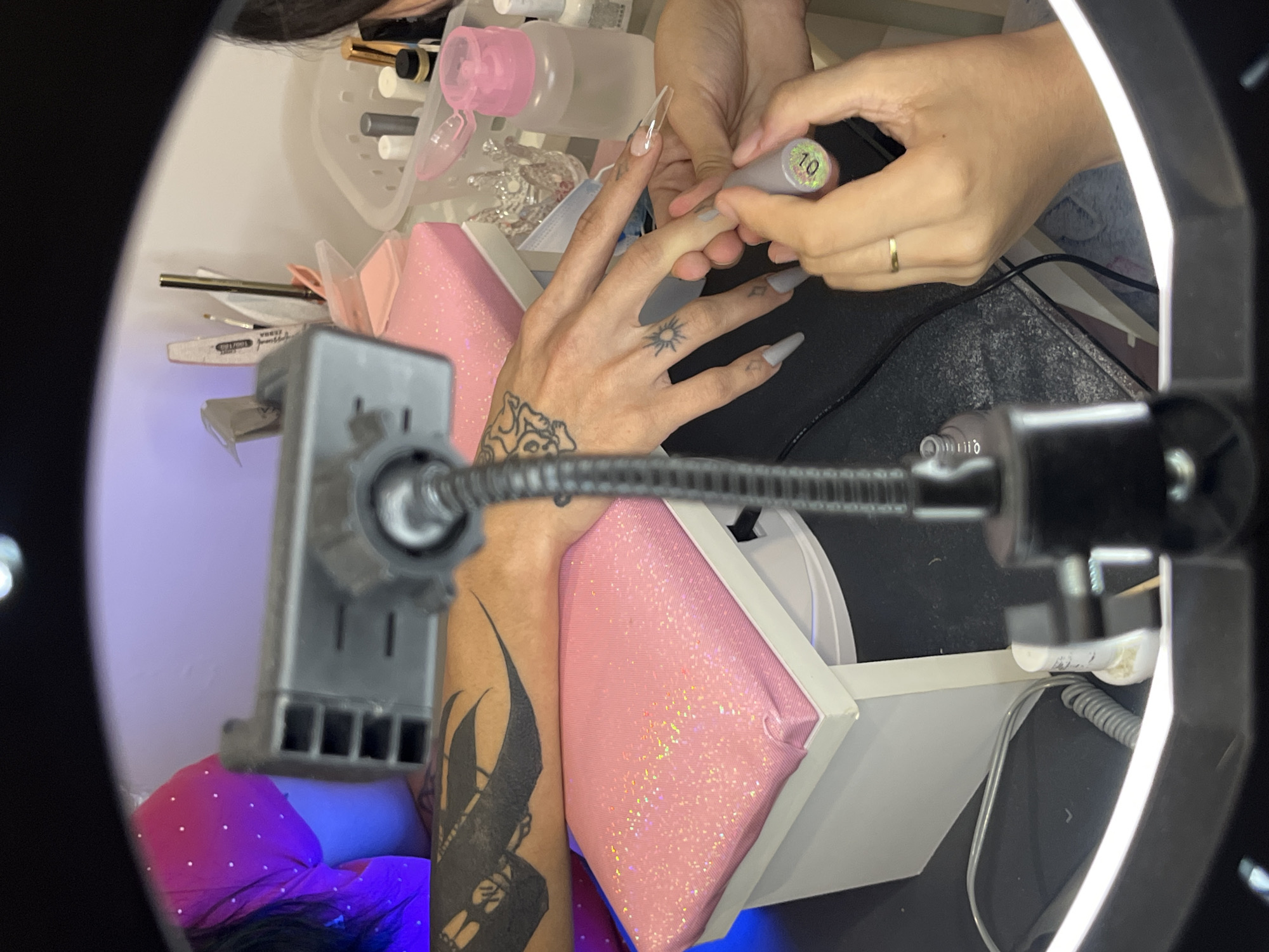 The image provides a top-down perspective of a nail service in progress, viewed through the center of a ring light. Two pairs of hands are the primary subjects. The client's left hand and forearm, extensively adorned with black tattoos including an intricate design on the forearm and a small sun on the back of the hand, are resting on a shimmering pink hand cushion. The client's nails are long and pointed; some are clear artificial extensions, while others are painted with a matte light grey or white polish.
The nail technician's hands are seen delicately applying polish from a silver bottle marked "10" to the client's nails. The technician wears a simple gold ring on one finger. Surrounding the workspace are various manicure tools and products: a pink pump-top liquid dispenser, multiple clear bottles, and a nail file visibly marked "100/180". A small face mask is also partially visible among the supplies.
In the foreground, a smartphone holder or camera mount is attached to the ring light, suggesting the process might be recorded or live-streamed. A purple glow from a UV/LED nail lamp is visible beneath the client's arm, indicating the use of gel polish that requires curing. The setting appears to be an organized nail station, possibly in a salon or a dedicated home setup, with white shelves and a black work surface. This detailed manicure session is taking place in Buenos Aires, Argentina.