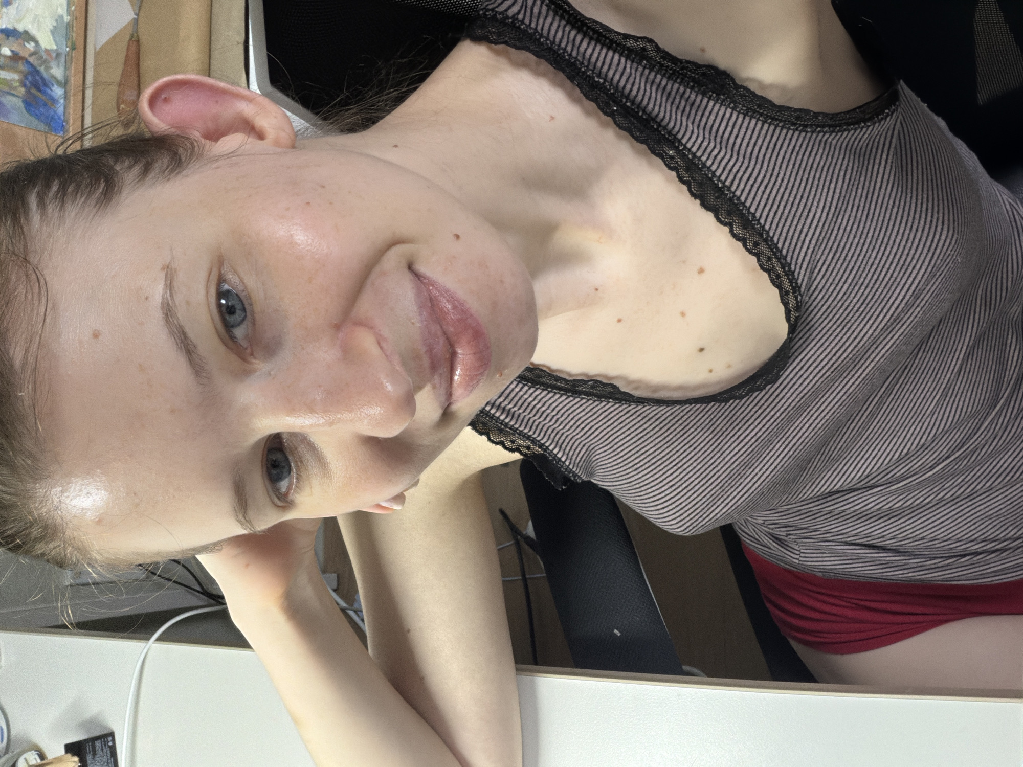 A young woman with fair skin and blue eyes is taking a selfie indoors. She is wearing a gray and black striped tank top with lace trim and red underwear. Her left arm is bent, and her hand is resting on her head. The background includes a desk, a chair, and some art supplies. The image appears to have been taken in Dongducheon-si, South Korea.