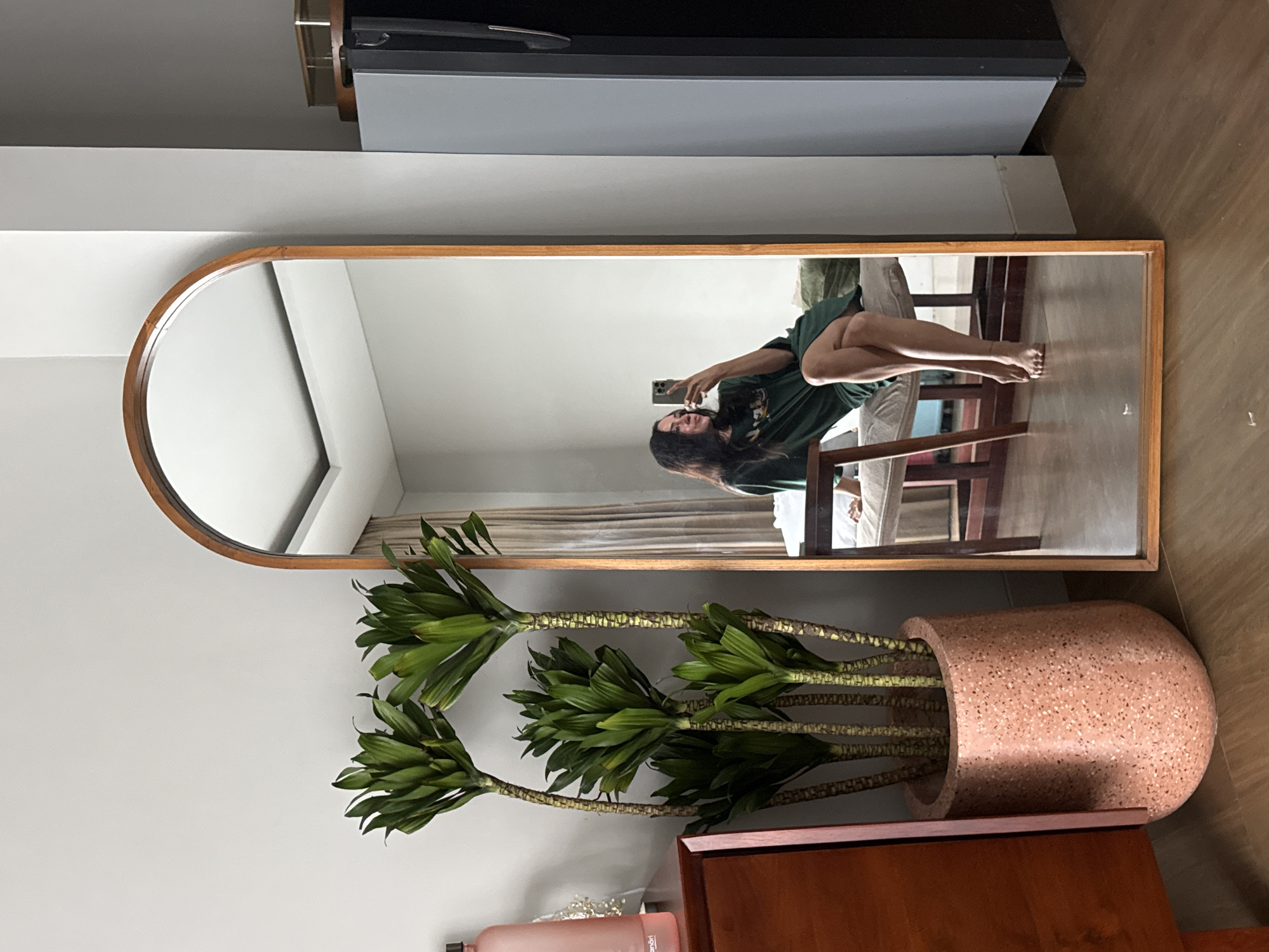 In North Kuta, Indonesia, a woman in a dark green dress is taking a selfie in a full-length arched mirror. She is seated on a bed or bench with her legs crossed, holding her phone up to capture her reflection. The mirror is framed in wood and stands on the floor, reflecting the room's interior. To the left of the mirror, a large potted plant with multiple stalks and green leaves is visible. The plant is in a textured, peach-colored pot. A portion of a wooden dresser or cabinet is also visible to the far left. The background includes a light-colored wall and curtains. To the right of the mirror, a tall, gray appliance, possibly a refrigerator, is present. The floor is made of light-colored wood.