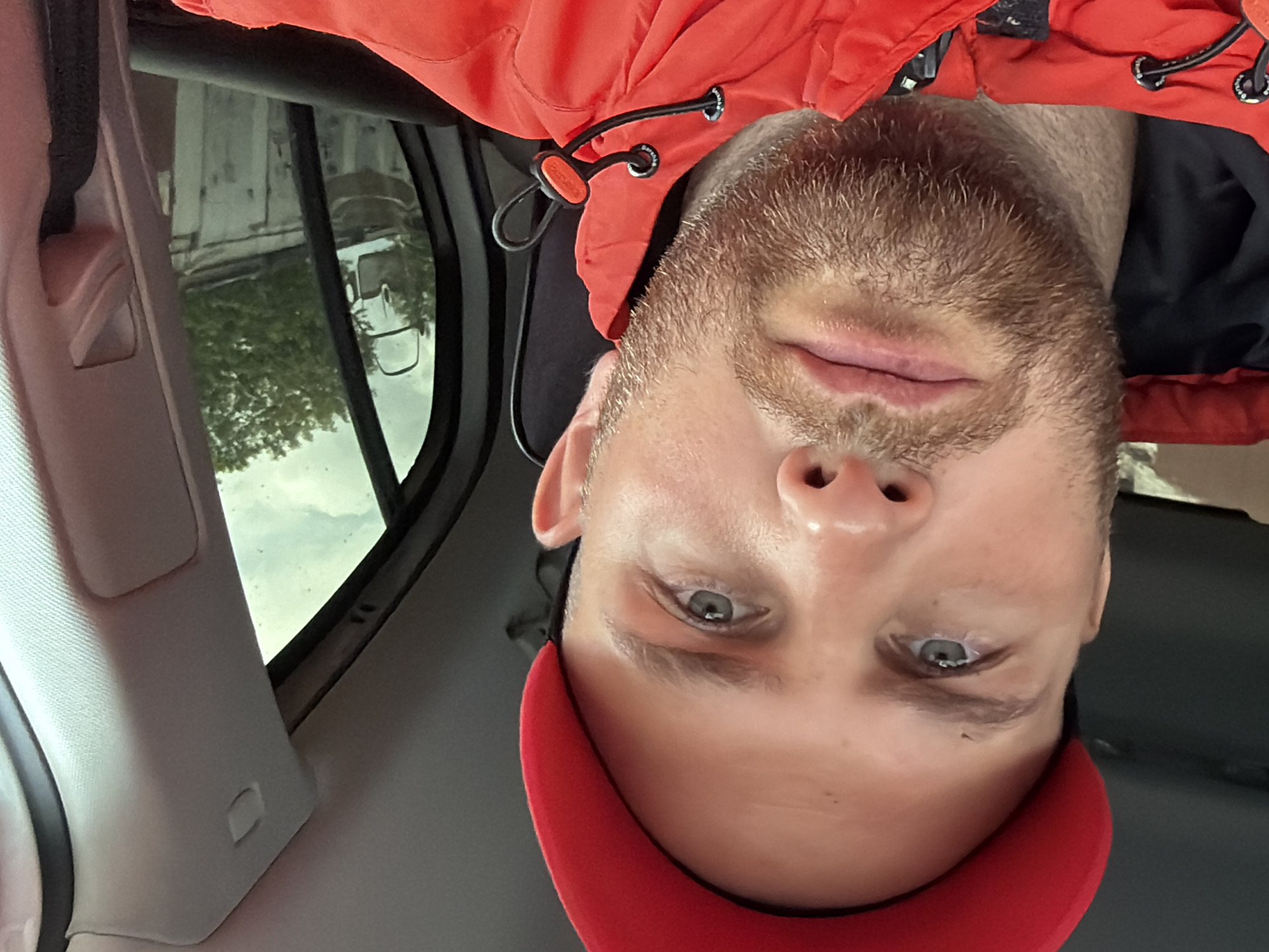 The primary image, originally presented inverted, captures a close-up selfie portrait of a man inside a car. The man, appearing to be in his 20s or 30s, has fair skin, light blue or grey eyes, and light brown stubble or a short beard. He looks directly at the camera with a neutral expression. He is wearing a vibrant red jacket, which appears to be a puffer or windbreaker style, with black inner lining visible near his neck. A black drawstring with a toggle, featuring the faint brand name "Bershka," is visible on the jacket's collar area. Below his chin, part of a red cap or hat is visible.
The setting is the interior of a vehicle, with sections of the cream-colored ceiling trim and the A-pillar visible on the left side of the frame. Through the car window on the left, an outdoor scene is reflected and partially visible. This reflection shows a light-colored building, green tree foliage, and what appears to be a white van or truck. The sky outside is overcast, suggesting a cloudy daytime environment without direct sunlight. The scene provides a glimpse into a moment of the man's day, situated in Chaikovsky, Russia.