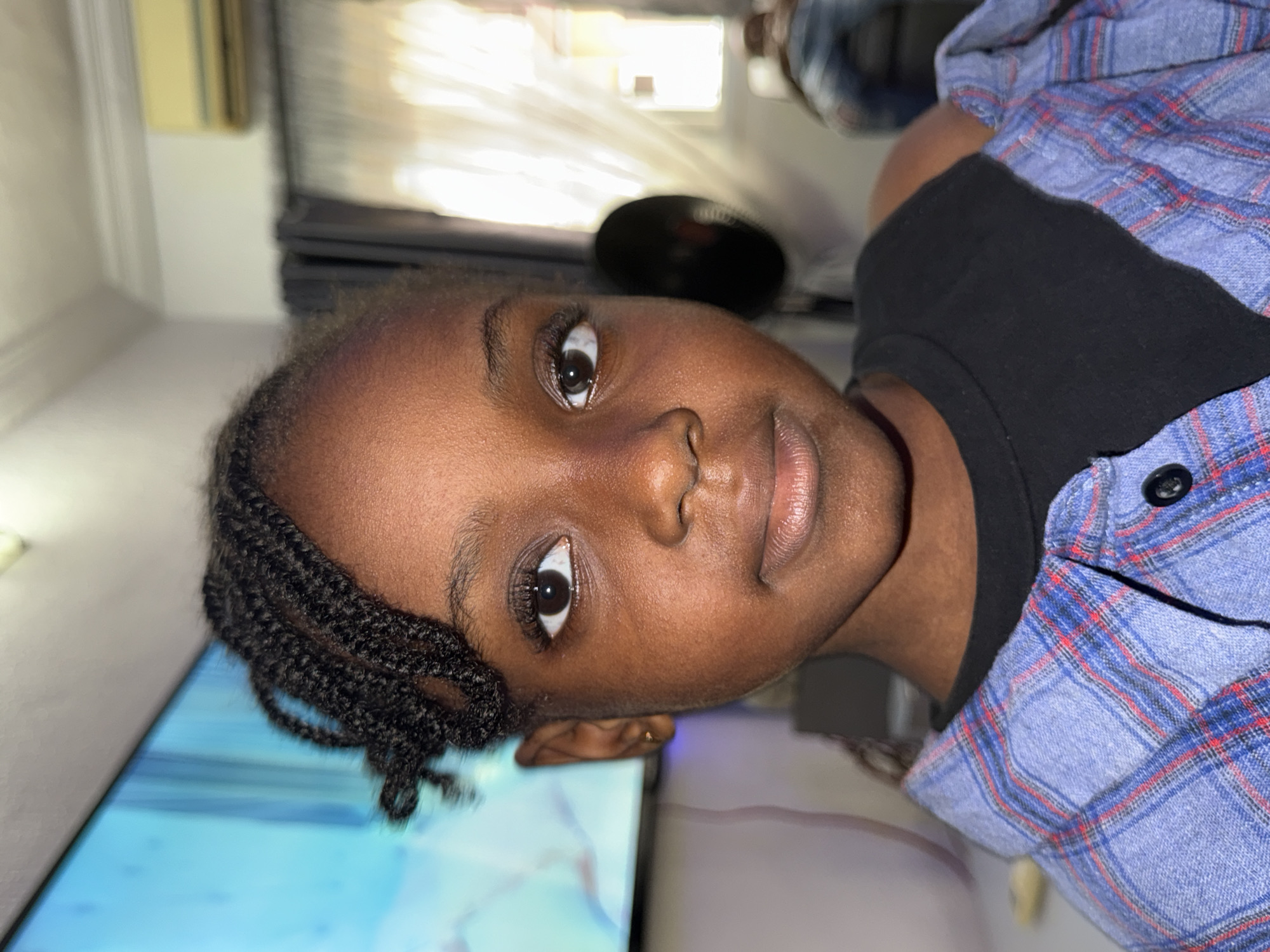 A close-up, portrait-style shot captures a young person, likely a child, with dark skin, looking directly at the camera with a neutral expression. Their head is tilted, suggesting the photo might have been taken from a slightly low angle or with a rotated camera. The subject has dark, neatly braided hair on the side of their head, and prominent dark eyes with a noticeable reflection of a light source in the pupils. They are wearing a black t-shirt beneath a blue, red, and white plaid button-up shirt.
The background is an indoor setting, appearing to be a home in Ado, Nigeria. To the subject's left (bottom of the image when oriented), a large, bright light blue surface is visible, which appears to be a television screen displaying abstract blue and white patterns. Behind the subject, the background is softly blurred, showing a light-colored wall, some indistinct dark furniture or electronics, and a bright, sunlit window or opening, indicating daylight outside. The lighting primarily illuminates the subject's face, highlighting their features. There is no visible text.
