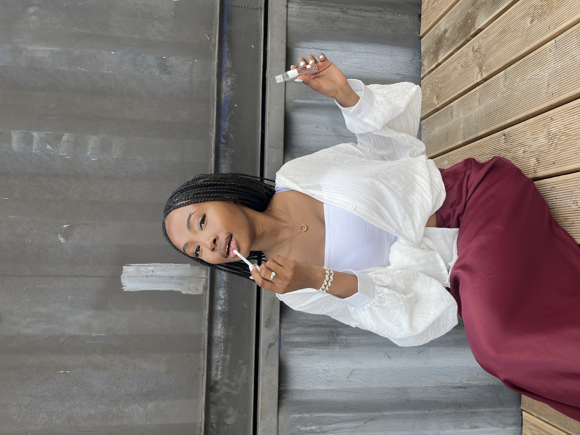 A young Black woman with braided hair is sitting on a wooden bench in front of a large, textured, grey industrial wall. She is wearing a white tube top, an oversized white open-front shirt with voluminous sleeves, and a flowy, burgundy-colored skirt. She is applying lip gloss to her lips with a small applicator and holding a clear tube of lip gloss in her right hand. She is wearing a gold pendant necklace and a beaded bracelet on her left wrist. The background wall shows signs of wear and tear, with scuffs and smudges. The wooden bench appears to be made of planks. The overall lighting suggests an outdoor setting, possibly during the daytime. There is no indication that this scene is located in Kazan, Russia.