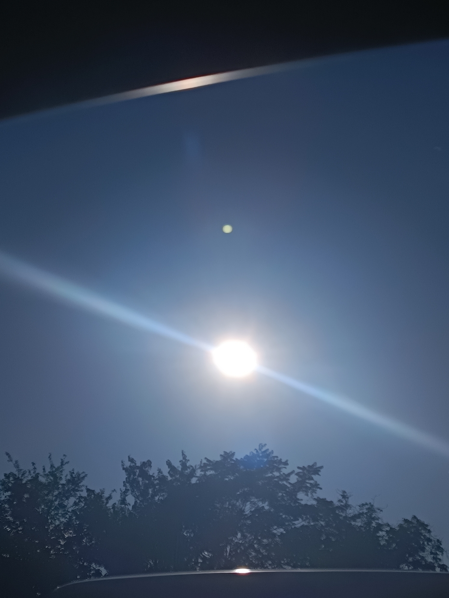 The image displays a bright sun in a hazy blue sky, creating a lens flare effect that extends diagonally across the frame. A smaller, yellowish orb is visible higher in the sky, potentially representing a celestial body or a photographic artifact. Silhouetted against the sky are the tops of dense, dark green trees and a dark horizontal structure, possibly a vehicle roof, at the bottom of the image. The overall scene is consistent with a daytime view of the sky from within the city of Zaporizhzhia, Ukraine, likely captured from a ground-level perspective.