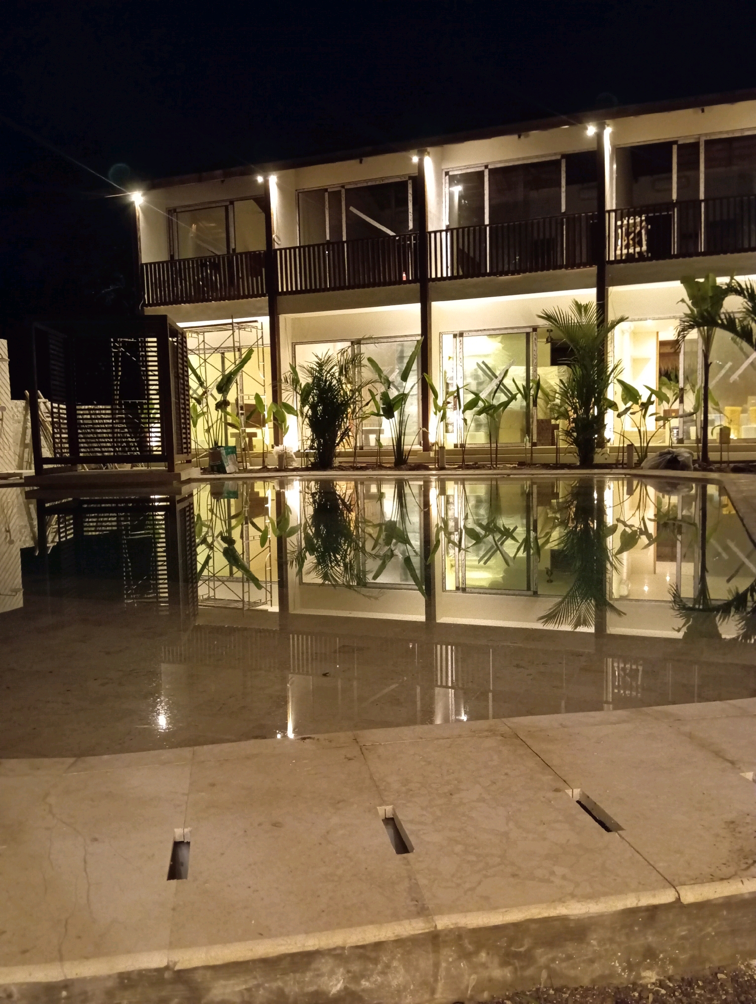 In Kuta, Indonesia, at night, a contemporary two-story building with a dark wooden railing on the second-floor balcony is illuminated by warm outdoor lighting. Large windows reflect the surrounding environment, and tropical plants are visible both inside and outside. A shallow, flooded area in the foreground acts as a mirror, reflecting the building, plants, and railing. Concrete slabs with rectangular cutouts are visible at the edge of the flooded area. The scene is dark, with the exception of the artificial lighting.