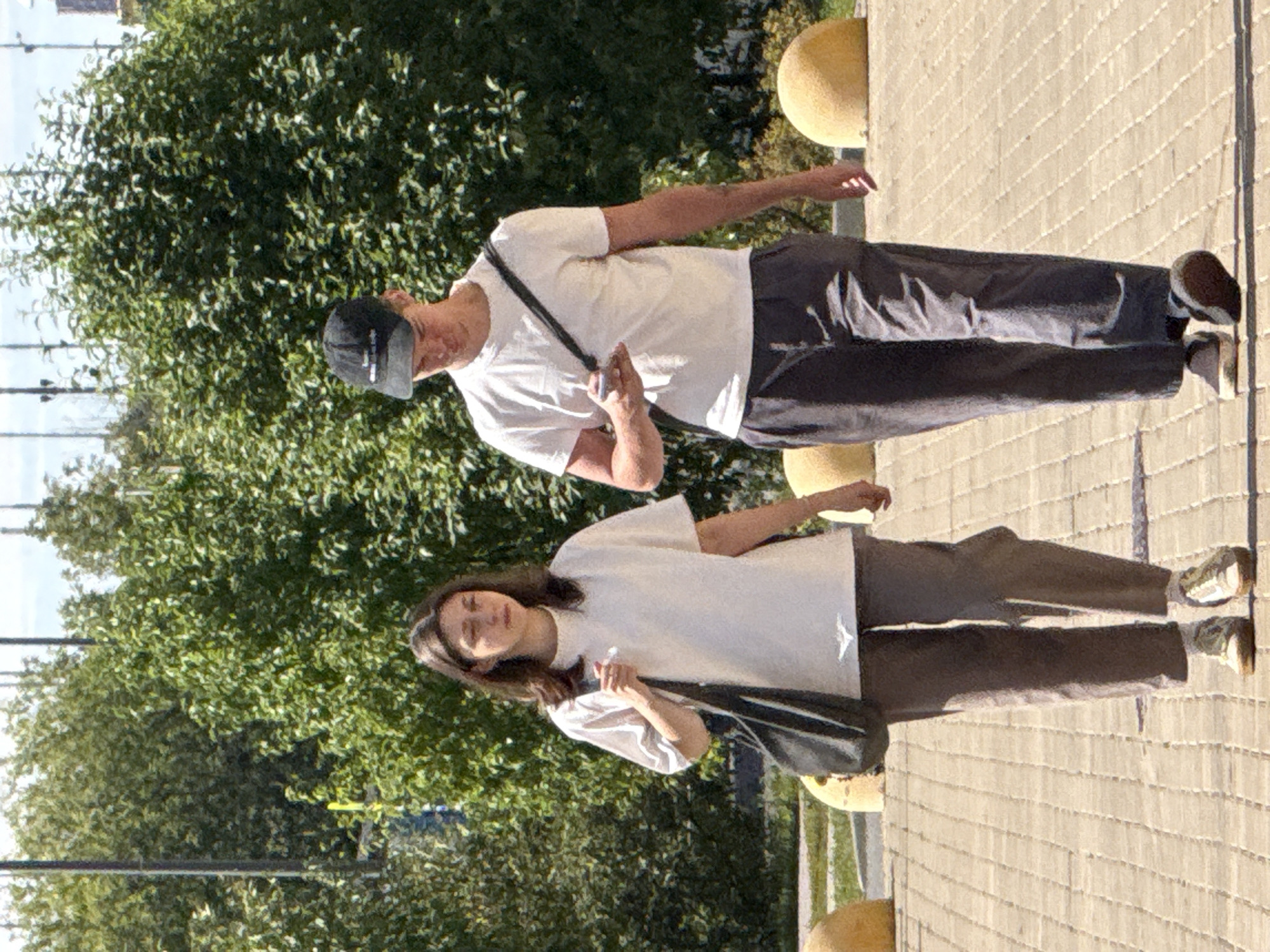 Two individuals, a young woman and a young man, are walking side-by-side on a paved walkway under daylight. The woman, on the left, has shoulder-length dark hair and is wearing a loose-fitting white t-shirt and grey pants. She carries a dark-colored bag over her left shoulder. The man, on the right, is wearing a white t-shirt, dark grey pants, and a black baseball cap. He has a black strap across his chest and is looking at his phone. The walkway appears to be made of light-colored, rectangular pavers. Behind them, dense green foliage forms a backdrop, with some parts of trees and other greenery visible. Two large, rounded, yellowish-orange bollards are positioned to the left and right of the walkway. The overall environment suggests an outdoor urban setting, possibly a park or a pedestrian area in Moscow, Russia, characterized by greenery and paved pathways.
