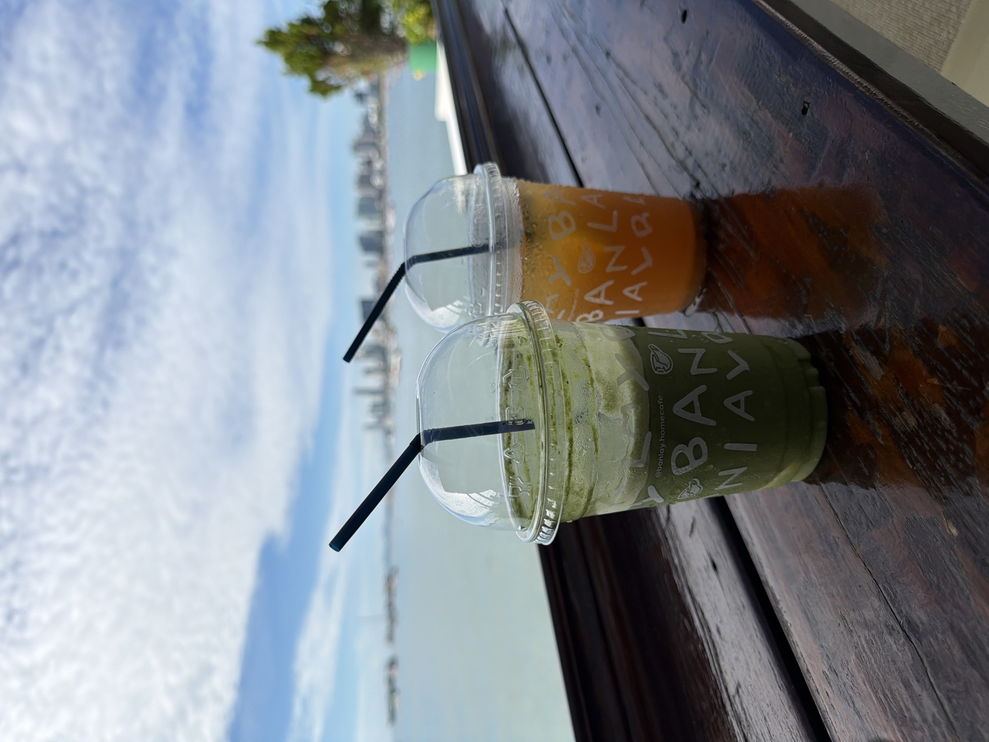 Two iced drinks, one green and one orange, sit on a wooden table with a view of the ocean and a city skyline in the background. The green drink is in the foreground on the left, with a clear plastic cup and lid, and a black straw. The cup has white text that reads "@banlay.homecafe BANLAY". The orange drink is to the right of the green drink, also in a clear plastic cup with a lid and a black straw. Its cup has white text that reads "BANLAY". The ocean is visible behind the drinks, with boats and a city skyline on the horizon. The sky is blue with white clouds. The overall scene suggests a relaxed setting, likely a cafe or restaurant with an outdoor seating area, overlooking the sea in Pattaya City, Thailand.