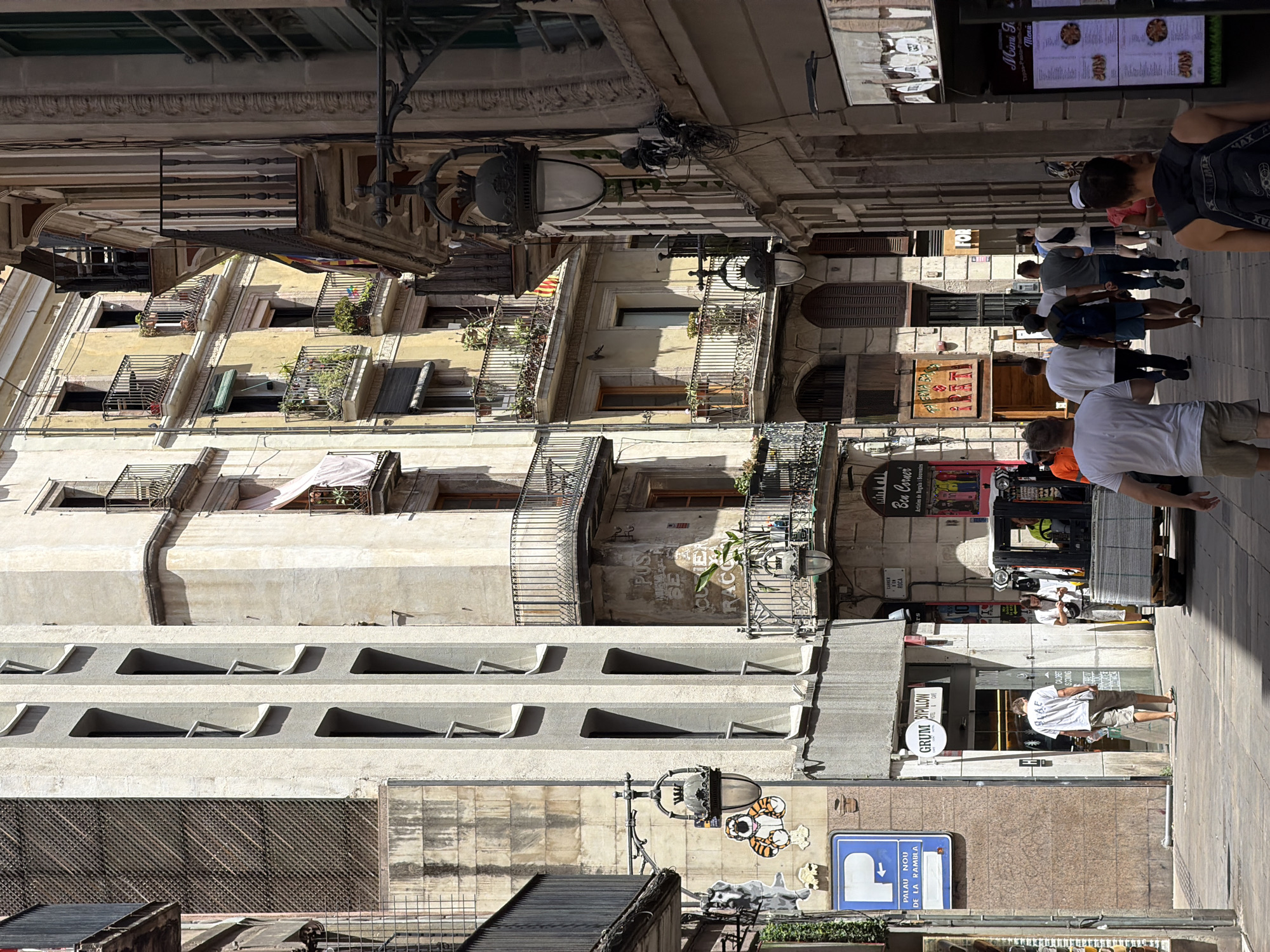 The image captures a street scene in the Gothic Quarter of Barcelona, Spain. The narrow street is flanked by old, tall buildings with balconies adorned with potted plants. Sunlight casts strong shadows, indicating a sunny day.
In the foreground, a man wearing a white t-shirt and beige shorts walks away from the camera. To his right, a forklift is parked, carrying stacks of what appear to be tiles or construction materials. Another man in a white shirt and light-colored pants stands near the forklift, looking at his phone.
Further down the street, a group of men are walking away from the camera. The buildings display various signs and architectural details, including decorative ironwork on the balconies and ornate street lamps. One building has a sign that reads "GRUM BYPILLOW MUSIC & CAFE." Another sign, partially visible, appears to be a menu for "Miami Food." A blue parking sign with a white 'P' and an arrow is mounted on the side of a building. Decorative decals of a dog and a tiger are affixed to the wall near the parking sign and a street lamp. The overall atmosphere is that of a bustling urban environment on a bright, sunny day.