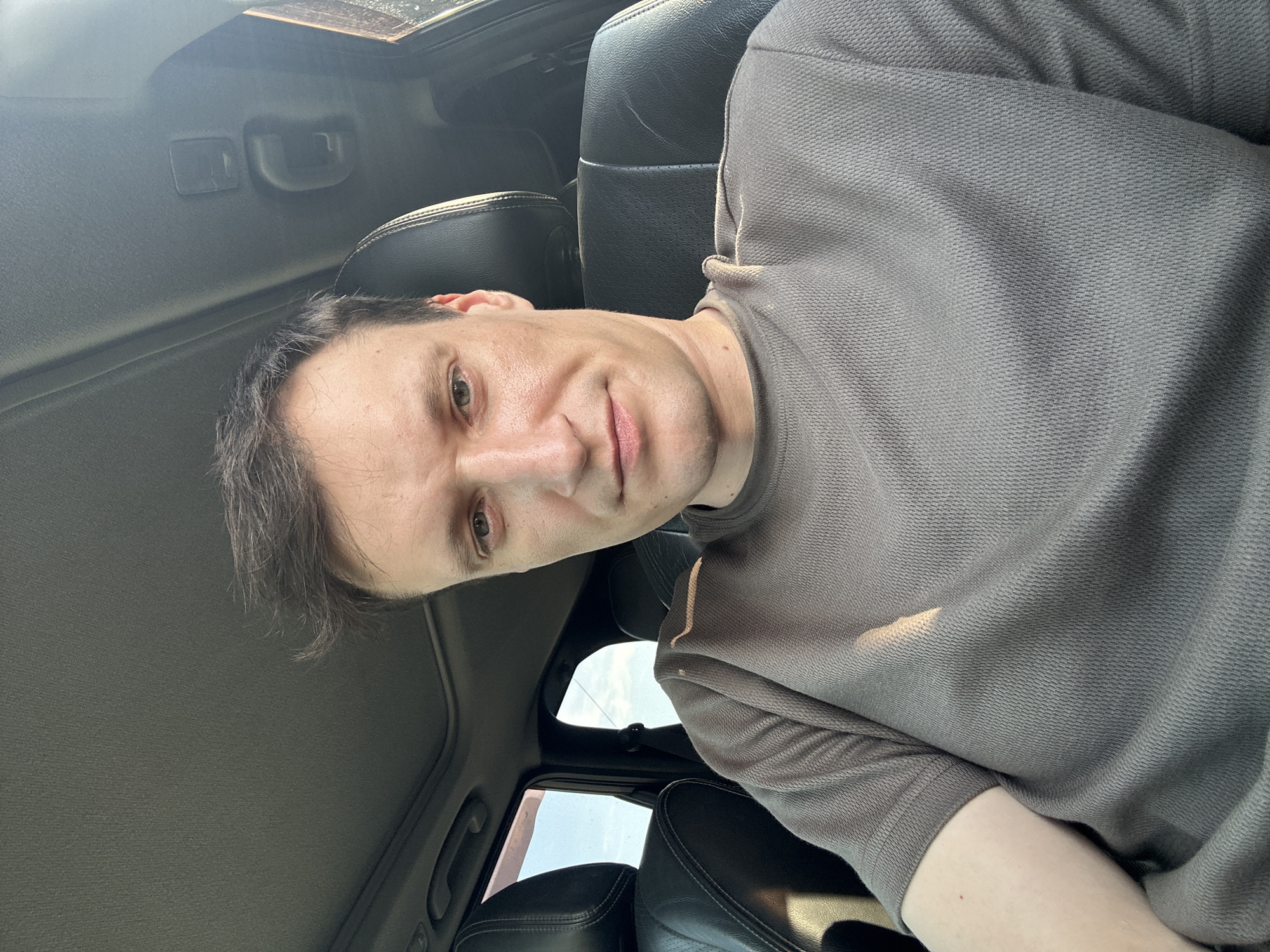 A close-up, slightly angled, medium shot captures a Caucasian male with short, dark brown hair and fair skin, seated in the driver's seat of a vehicle. The man is looking directly at the camera with a neutral expression. He is wearing a plain, taupe-colored t-shirt with a textured knit. The interior of the car is visible, featuring black leather seats and a dark gray headliner with a sunroof. Sunlight streams in from the right, casting a shadow across the man's right shoulder and shirt. The background shows a bright, hazy sky through the car windows, suggesting an outdoor setting during daylight hours. There are no discernible landmarks or elements that would indicate the specific location of Shatava, Ukraine.