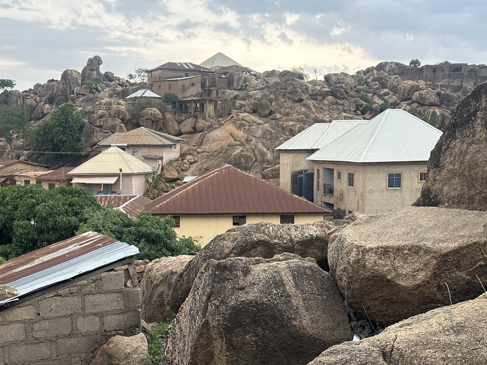 The image depicts a landscape in Jos, Nigeria, characterized by rocky terrain and dwellings constructed amongst the rock formations. The sky is overcast. The architecture consists of a mix of single and multi-story buildings, some with corrugated metal roofs of various colors. The buildings appear to be constructed from concrete blocks. The foreground is dominated by large, weathered boulders. Vegetation, including trees, is interspersed amongst the rocks and buildings. There are no people visible in the scene.