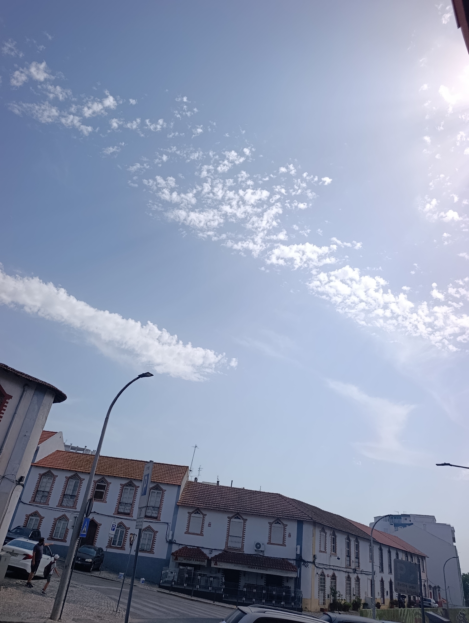 A street scene in Setúbal, Portugal, captured on a bright, sunny day. The sky is a vibrant blue with scattered white clouds, with the sun prominently positioned in the upper right corner, creating a lens flare effect. In the foreground, a street with cobblestone pavement runs diagonally across the image. Several cars are parked on the side of the street.
Dominating the middle ground are a series of traditional Portuguese buildings with white facades, reddish-brown tiled roofs, and arched windows featuring decorative brickwork. Some of these buildings appear to be shops or restaurants, with signage visible, including "RESTAURANTE VERDE E BRANCO". A modern street lamp with a curved pole stands prominently in the left-center of the frame.
On the sidewalk in front of one of the buildings, two individuals, one adult and one child, are visible. The adult is wearing dark clothing and the child is wearing a light-colored shirt and shorts. They appear to be walking or standing near a white car. The scene depicts a typical urban environment with a mix of traditional architecture and everyday street life.