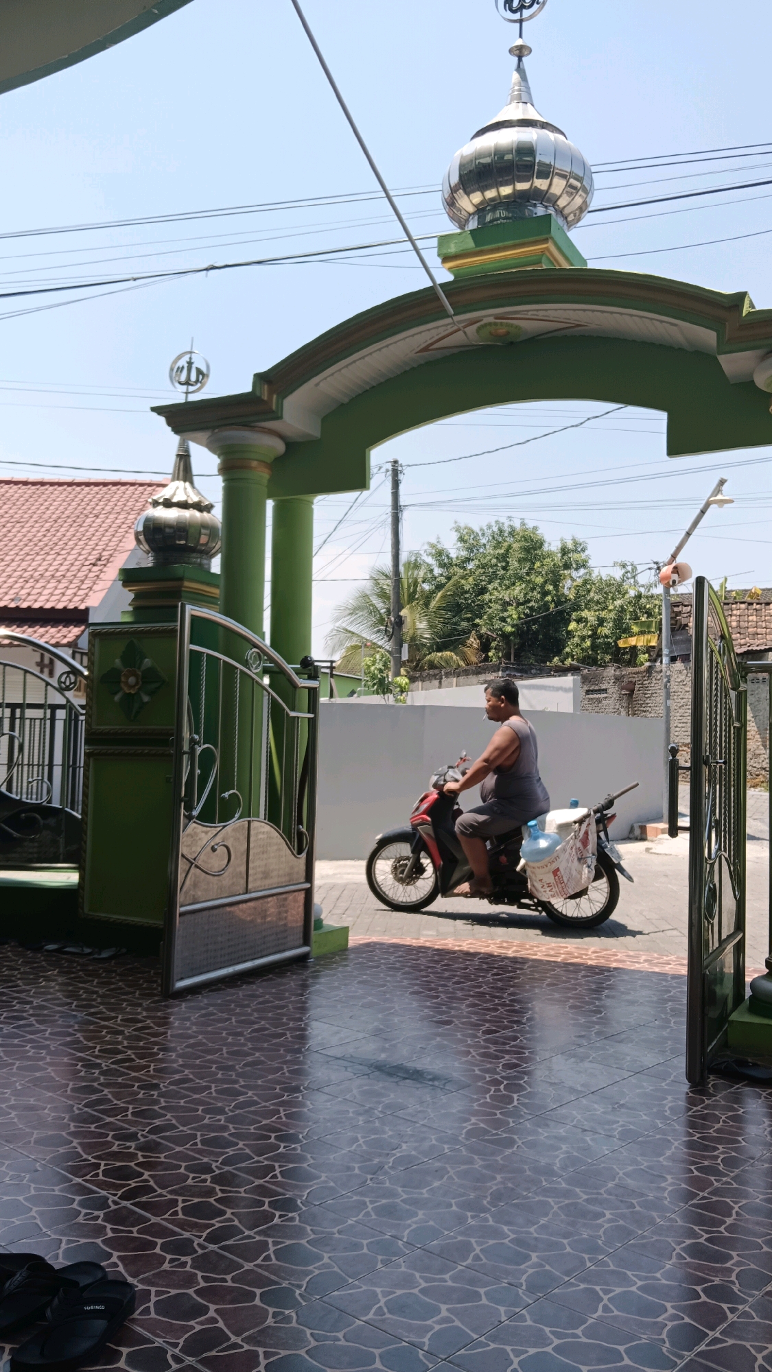 A man rides a red and black motorcycle through an archway with green pillars and a decorative metallic dome. He wears a grey tank top and shorts, and his motorcycle is equipped with a large white plastic container and a white bag strapped to the rear. The motorcycle is in motion, with its front wheel turned slightly to the left. The ground is tiled with a patterned design. To the left of the archway is a green gate with metallic embellishments. In the background, there are houses with red roofs and lush green trees. The sky is clear and blue. The scene appears to be an entrance to a property or a religious building in a sunny, outdoor environment, possibly in the city of Ngawi, Indonesia, given the architectural style and vegetation.