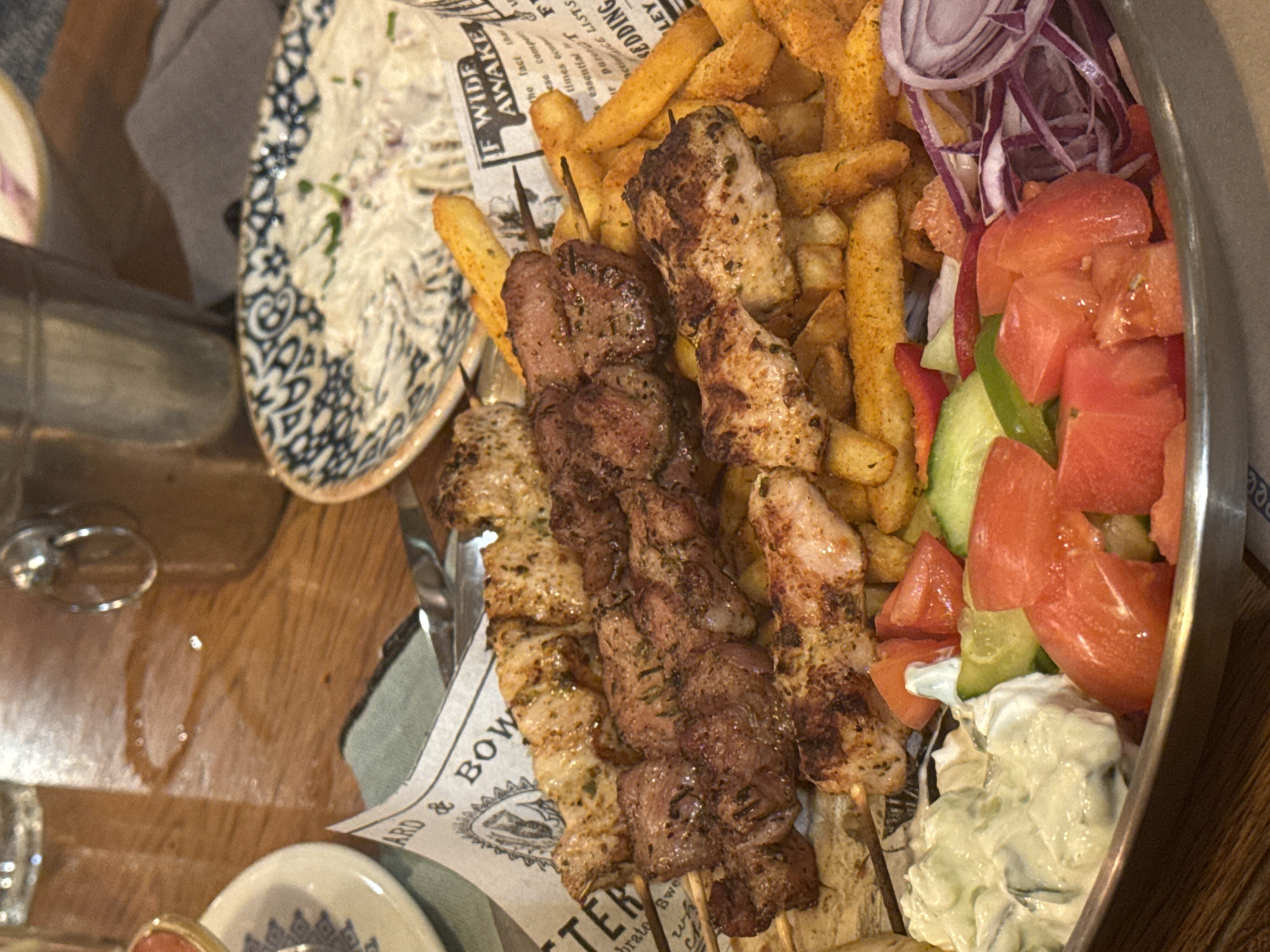 The image shows a close-up of a platter of food, likely in a restaurant setting. The platter contains souvlaki skewers (grilled meat on sticks), French fries, chopped tomatoes, cucumbers, red onions, a white sauce (possibly tzatziki), and some red bell pepper. The food is arranged on what appears to be a decorative paper liner, which has text and illustrations in English. The table beneath the platter is wooden. There are also blurred images of a water glass, a small plate with a blue design, and another dish with what looks like white sauce or salad.
There is no specific information in the photo that allows for determination of the location in the city of Varna, Bulgaria.