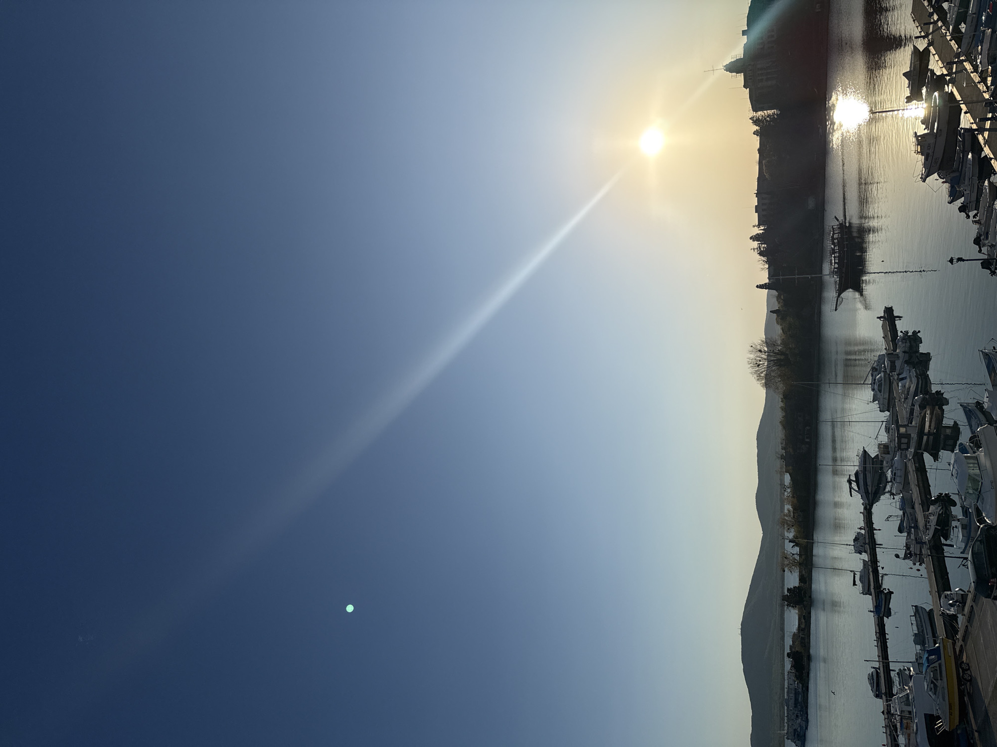 The image shows a waterfront scene in Sozopol, Bulgaria. The sky is a gradient from dark blue to light blue near the horizon, with the sun visible. Light from the sun reflects on the water. Boats are docked along a pier, and there's a building and hills in the background.