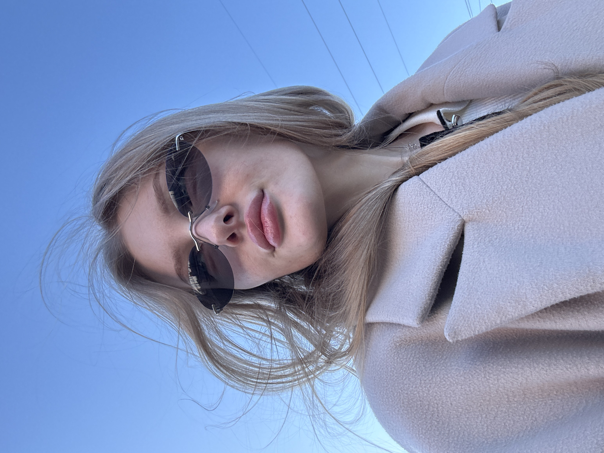 A young woman with blonde hair is seen from a low angle, wearing sunglasses and a light-colored coat. The background is a clear blue sky with power lines visible. The location is not specified as Perm, Russia, so no specific location details can be given.