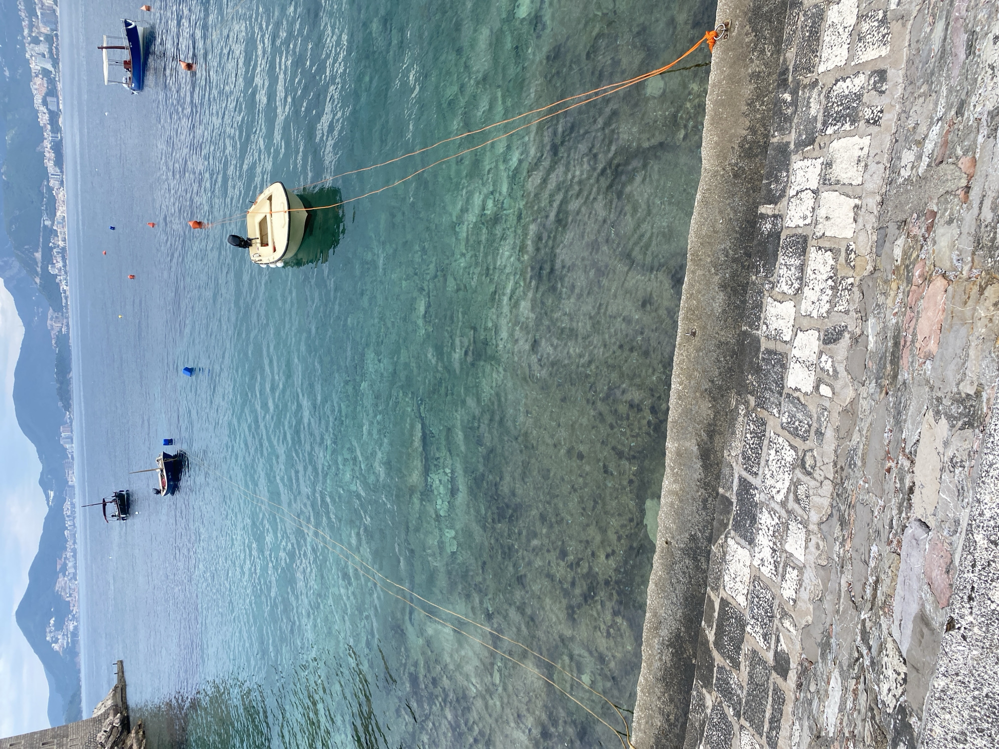 The image shows a view of the Adriatic Sea in Sveti Stefan, Montenegro. A small, light-colored boat is moored in the clear, turquoise water. Two orange ropes connect the boat to the stone quay on the right side of the frame. Further out in the water, a few other boats can be seen in the distance, along with buoys or markers. In the background, the coastline is visible with mountains and buildings. The stone quay is constructed from light and dark colored stone blocks with a concrete border.