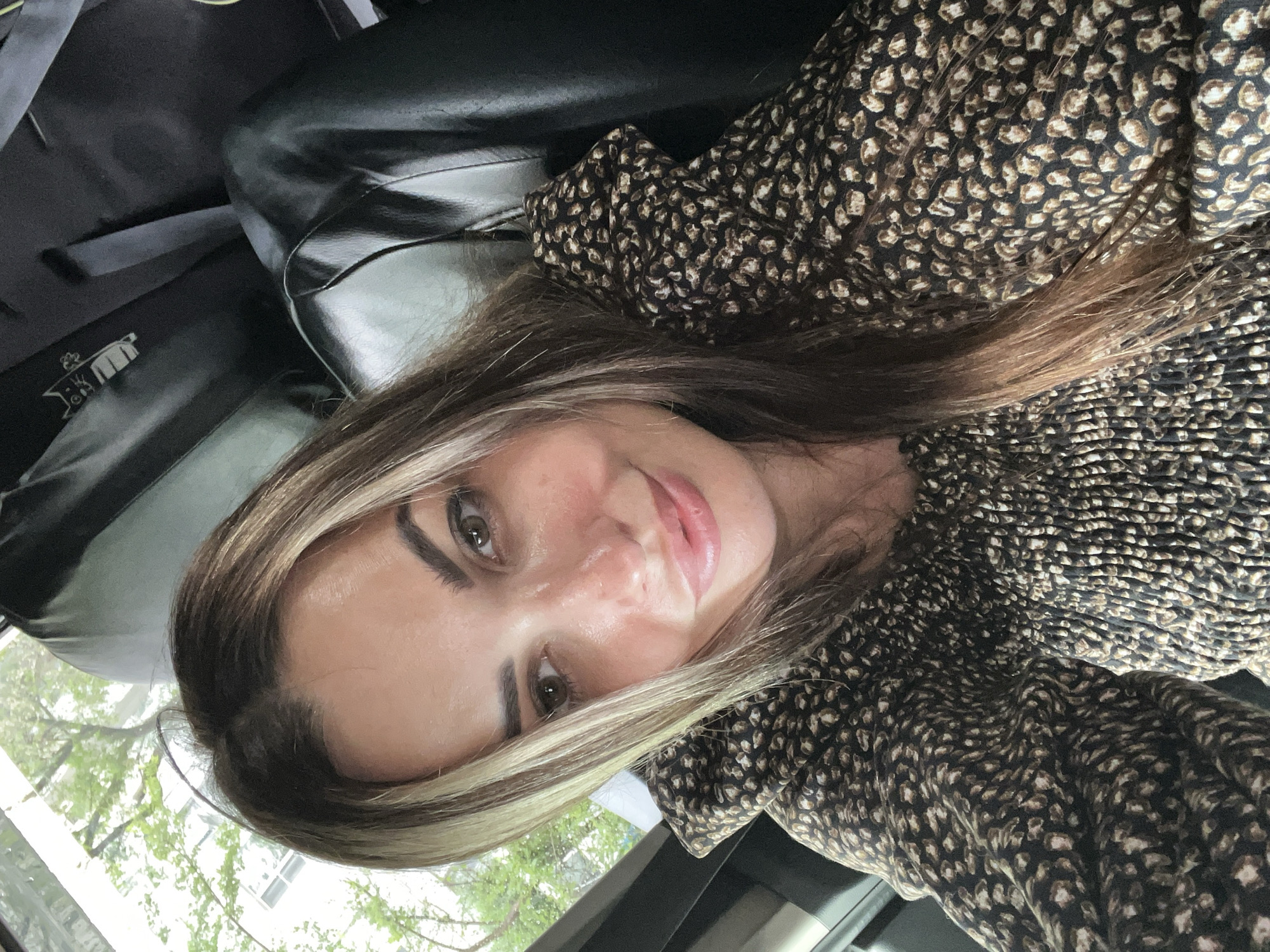 A woman with long, layered brown and blonde hair is looking at the camera and smiling slightly. She is wearing a smocked, long-sleeved top with a brown and tan animal print. Her head is tilted slightly to her left. The background is dark and appears to be the interior of a vehicle. A window with green foliage is visible to the left of the woman.