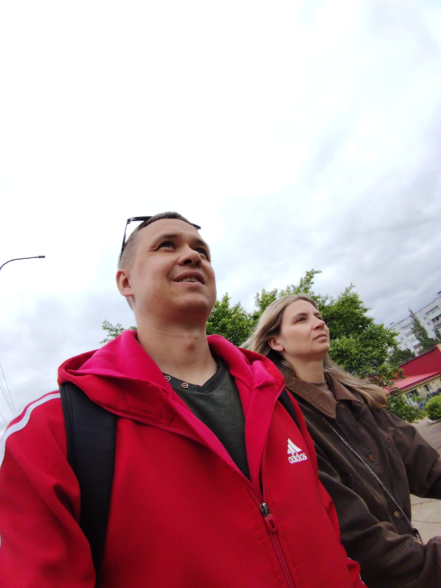 Two individuals, a man and a woman, are captured in a low-angle outdoor shot. The man, positioned closer to the camera, wears a red Adidas hooded jacket over a dark green shirt. Sunglasses are perched on his head. He is looking upwards and to the right. To his right, the woman has light-brown hair and wears a brown jacket. She also is looking in the same direction as the man. Behind them, green foliage is visible, along with a building with a red roof. In the background, an apartment building rises against an overcast sky. A street light is visible to the left of the man. The location is likely Chaikovsky, Russia.